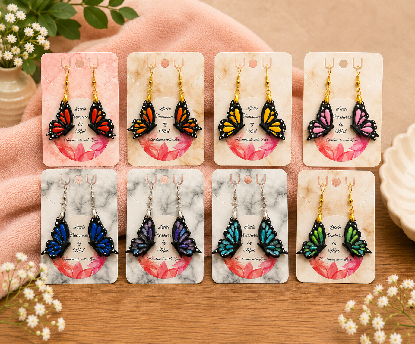 Colorful butterfly-shaped earrings displayed on cards with floral and marble backgrounds, arranged on a wooden surface with small white flowers in the corners.