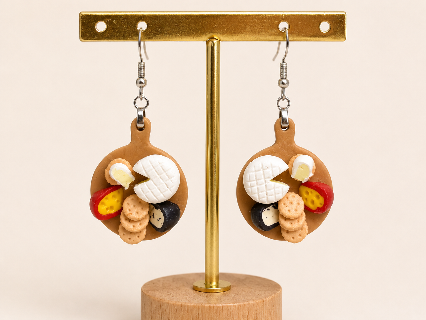 Earrings designed as miniature cheese and cracker platters with various types of cheese, crackers, and sausage on a display stand.