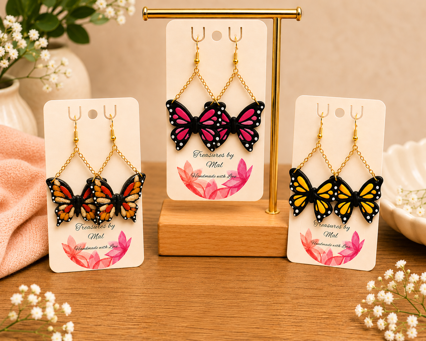 Three pairs of colorful butterfly earrings displayed on white cards with pink and red floral designs, set on a wooden surface with a small gold display stand, surrounded by white flowers and a pink cloth.
