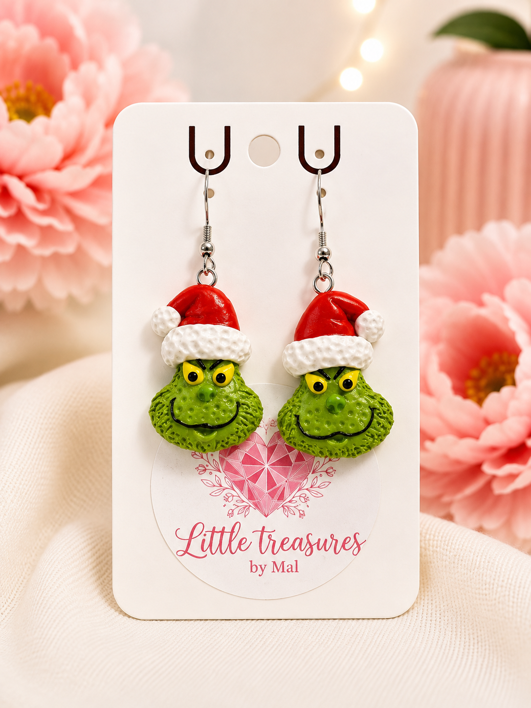 Green Grinch face earrings with red Santa hats on white card, pink flowers and soft background.