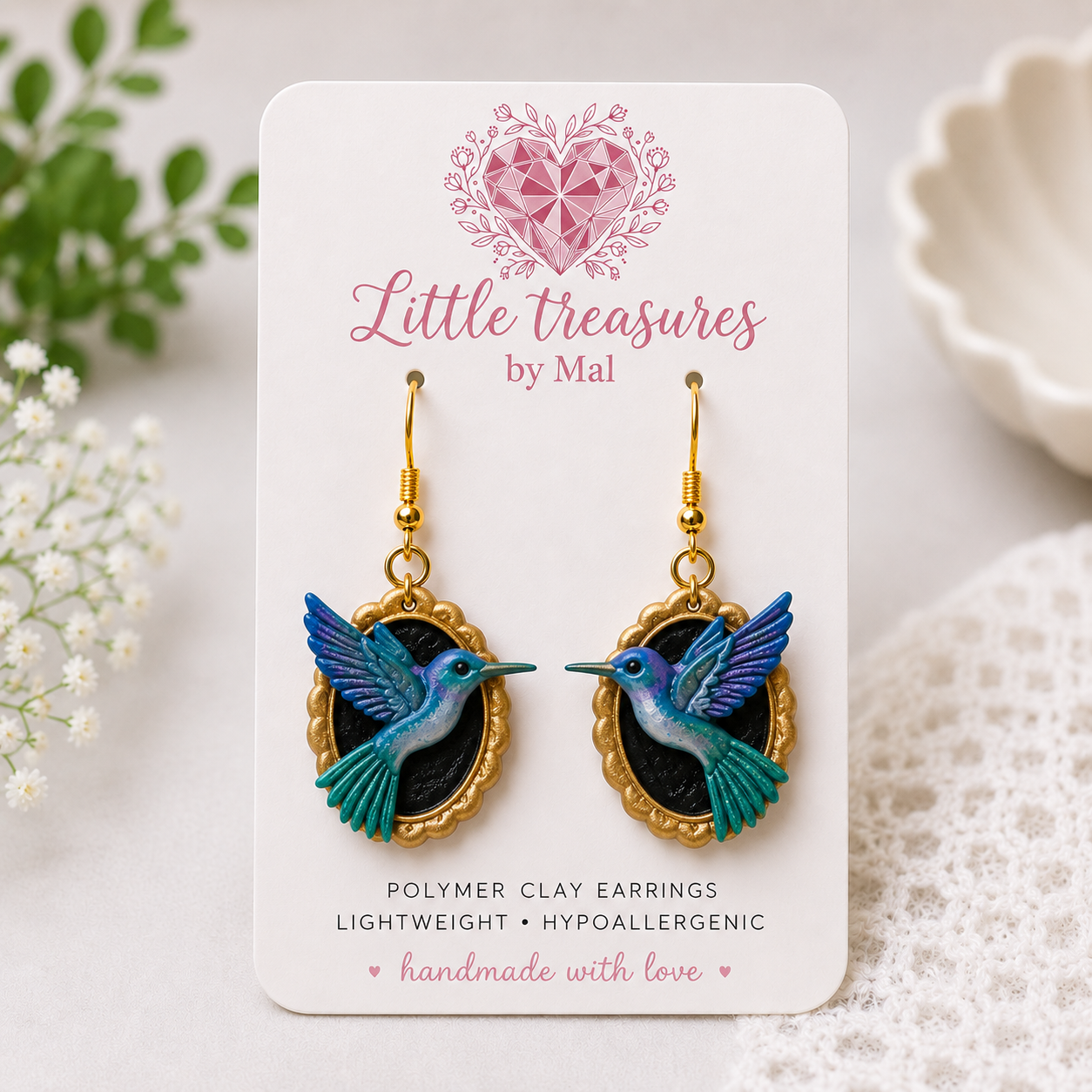 Colorful hummingbird earrings on a jewelry display card that reads "Little treasures by Mal." The earrings are gold with hummingbird charms featuring blue, purple, and green hues, and are lightweight and hypoallergenic.