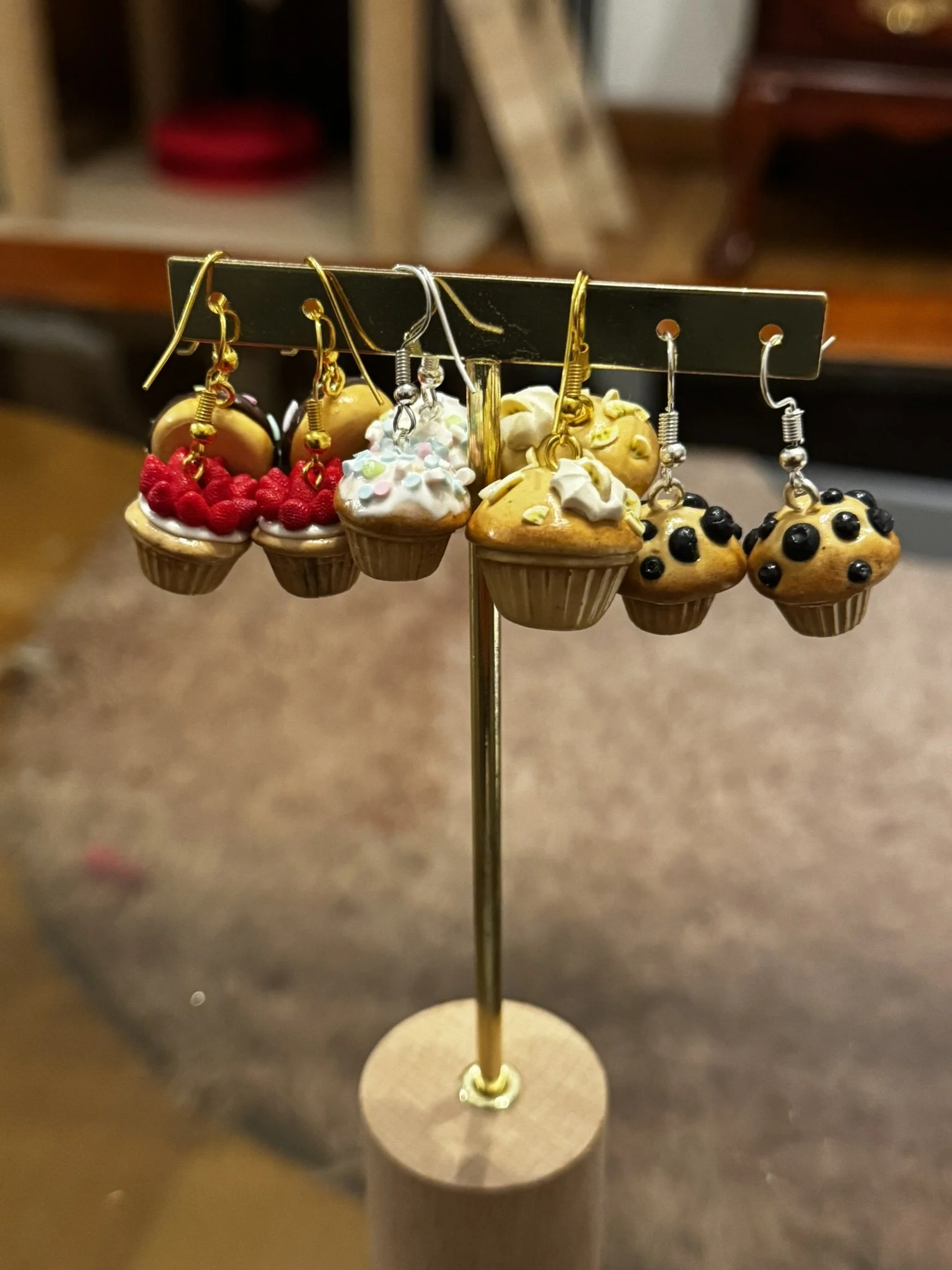 Miniature cupcakes with various toppings such as berries and sprinkles are displayed on a gold stand with hooks for earrings, holding six pairs of earrings.