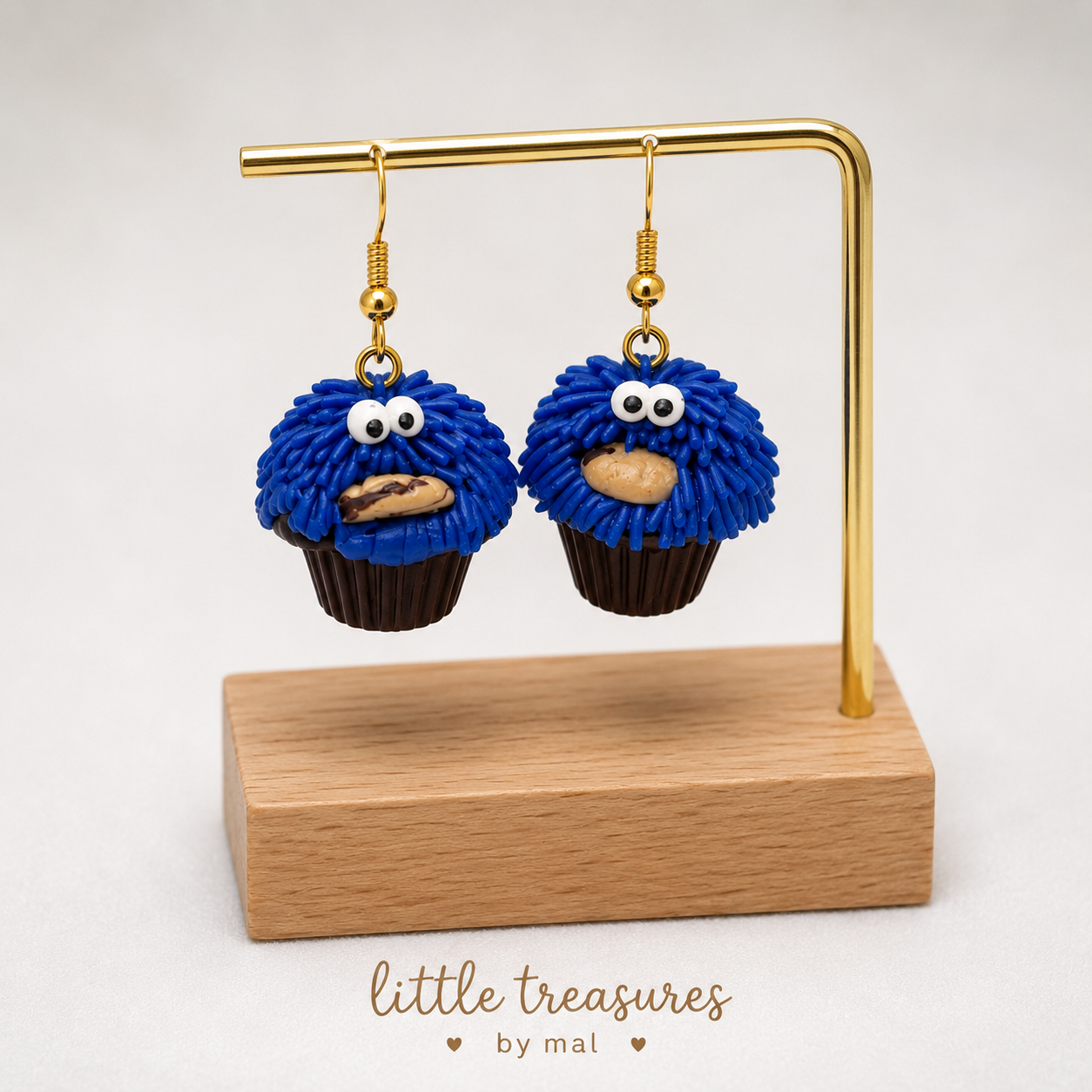 Earrings shaped like Cookie Monster with blue fringe fur and googly eyes, each holding a cookie, displayed on a stand with wooden base and labeled 'little treasures by mal'.