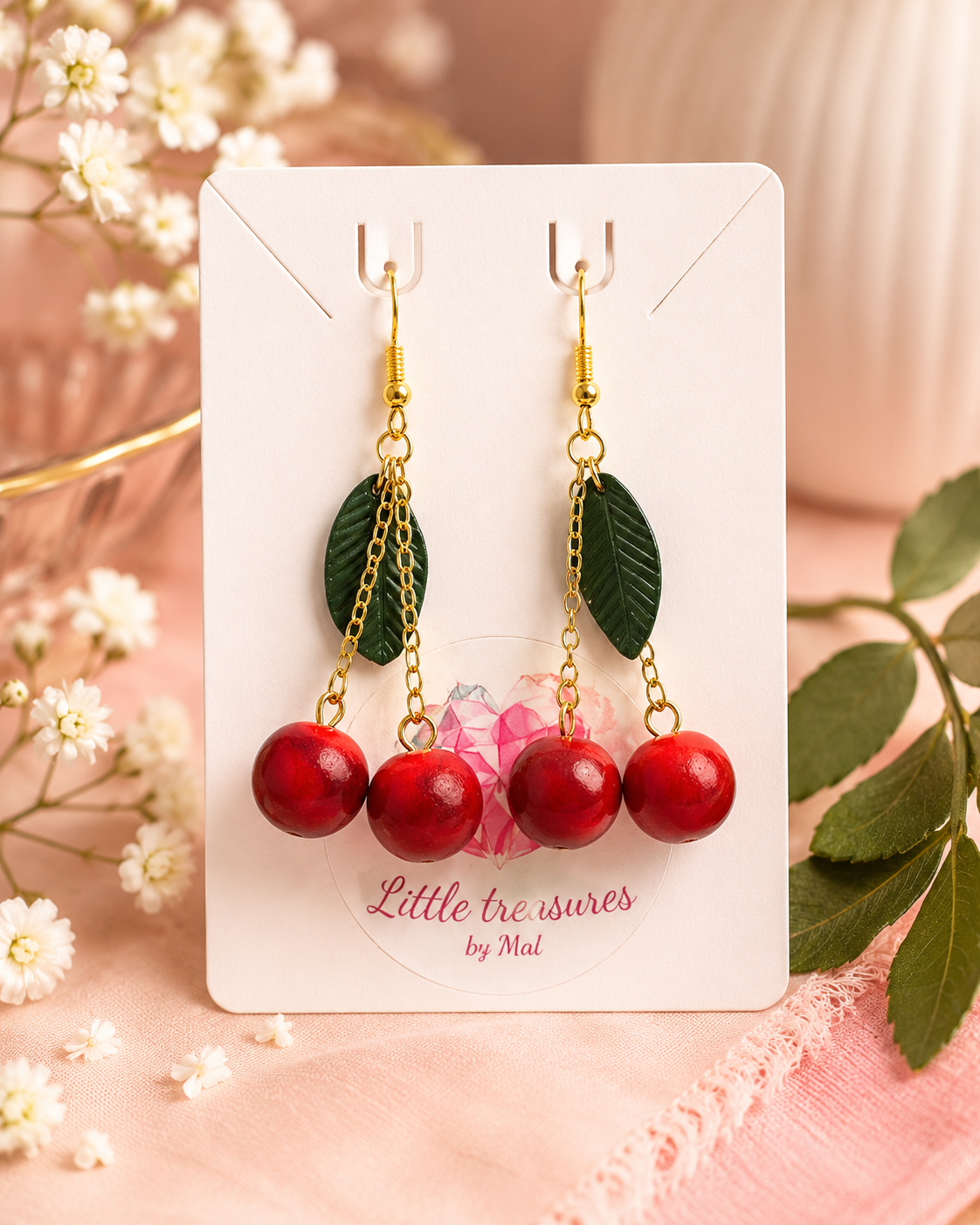 Pair of cherry-themed dangle earrings with green leaves and red cherry beads on a jewelry card labeled 'Little treasures by Mal', surrounded by white flowers and green leaves.