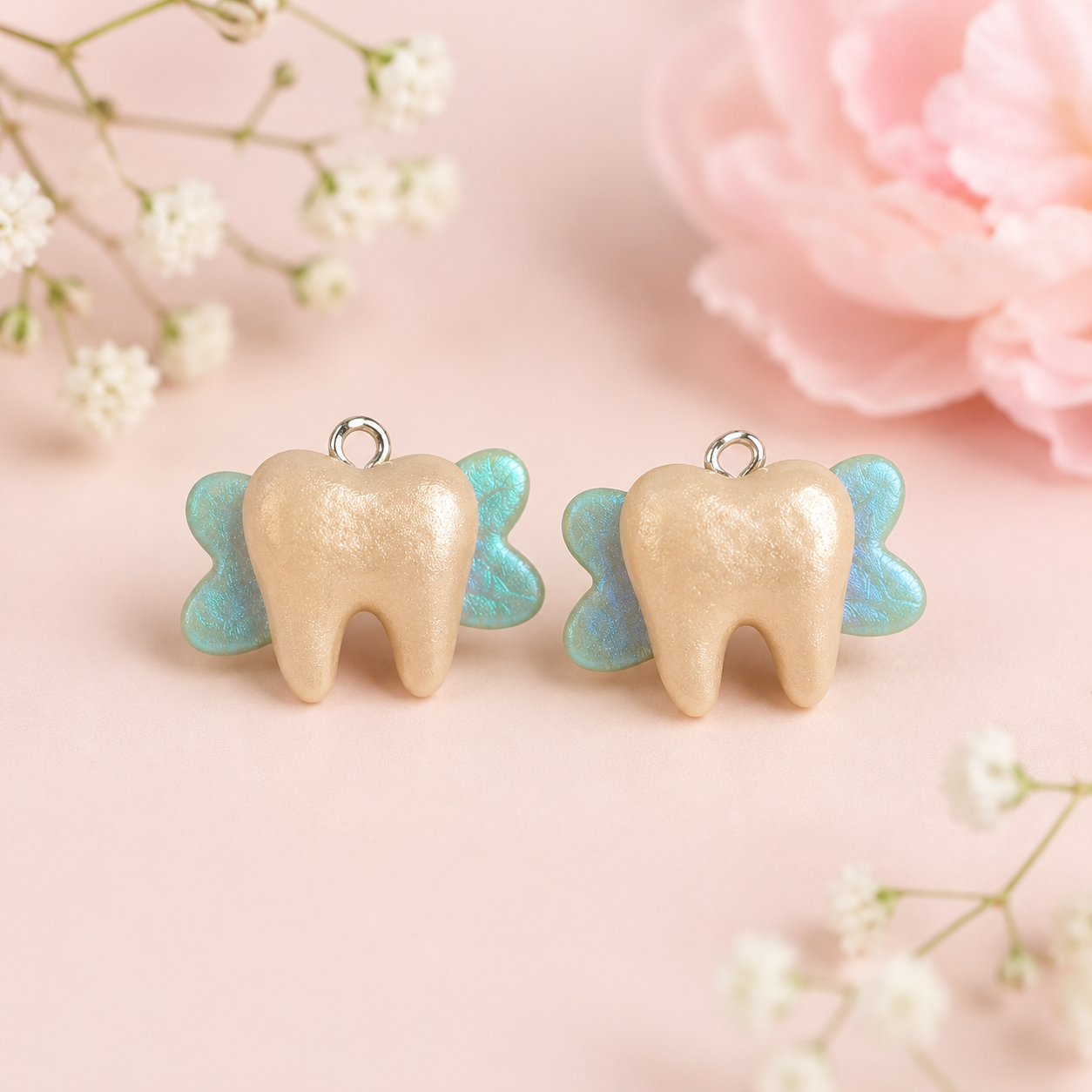 Two tooth-shaped charms with blue wings, placed on a pink background amid white flowers and a pink peony.