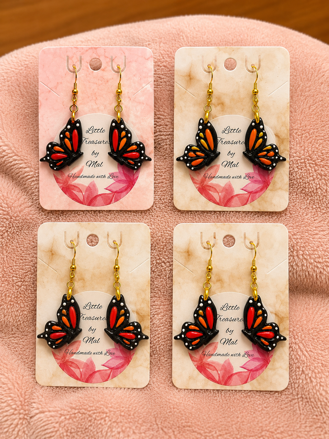 Set of four butterfly-themed earrings with black bodies and orange and white details, hanging on branded cards, displayed on a pink textured surface.