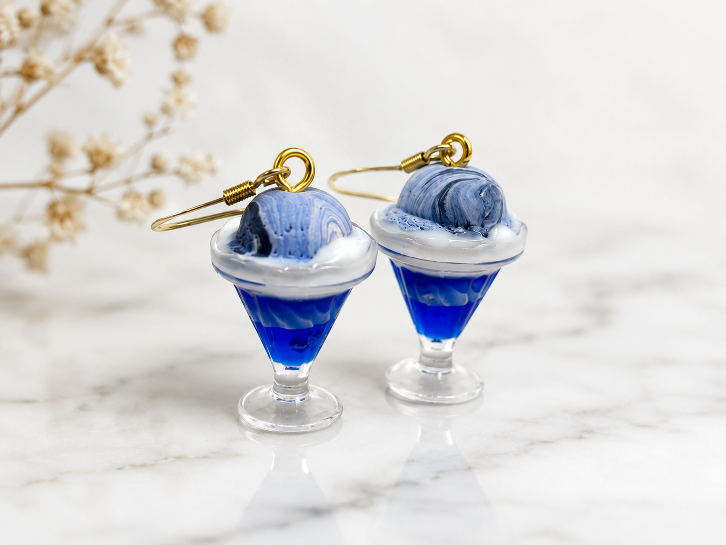 Earrings shaped like ice cream sundaes with blue and white swirled ice cream and blue glass bases on a white marble surface.