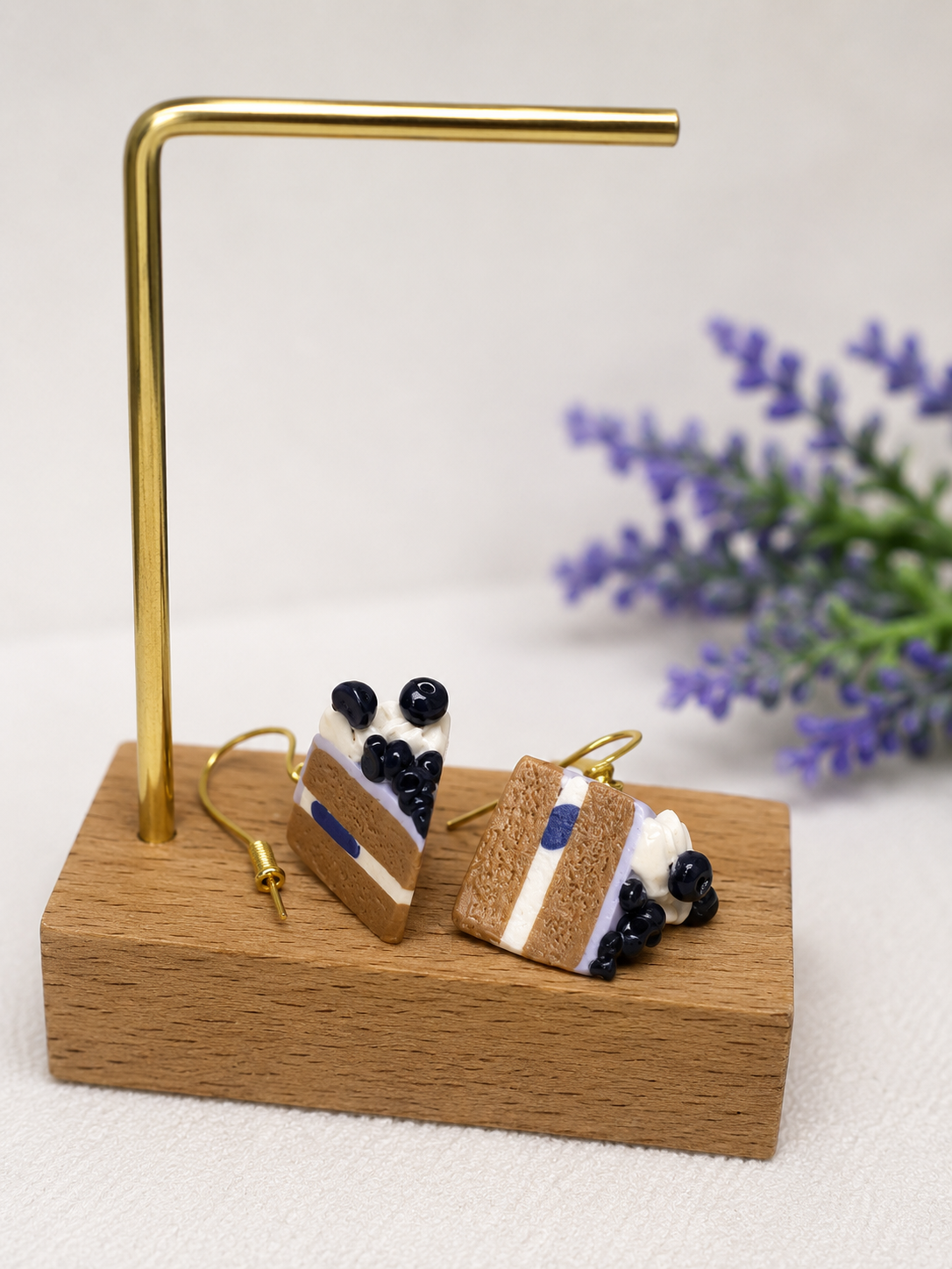 Dangle earrings designed to look like layered cake slices with whipped cream and blueberries, displayed on a wooden platform with a gold metal stand and purple flowers in the background.