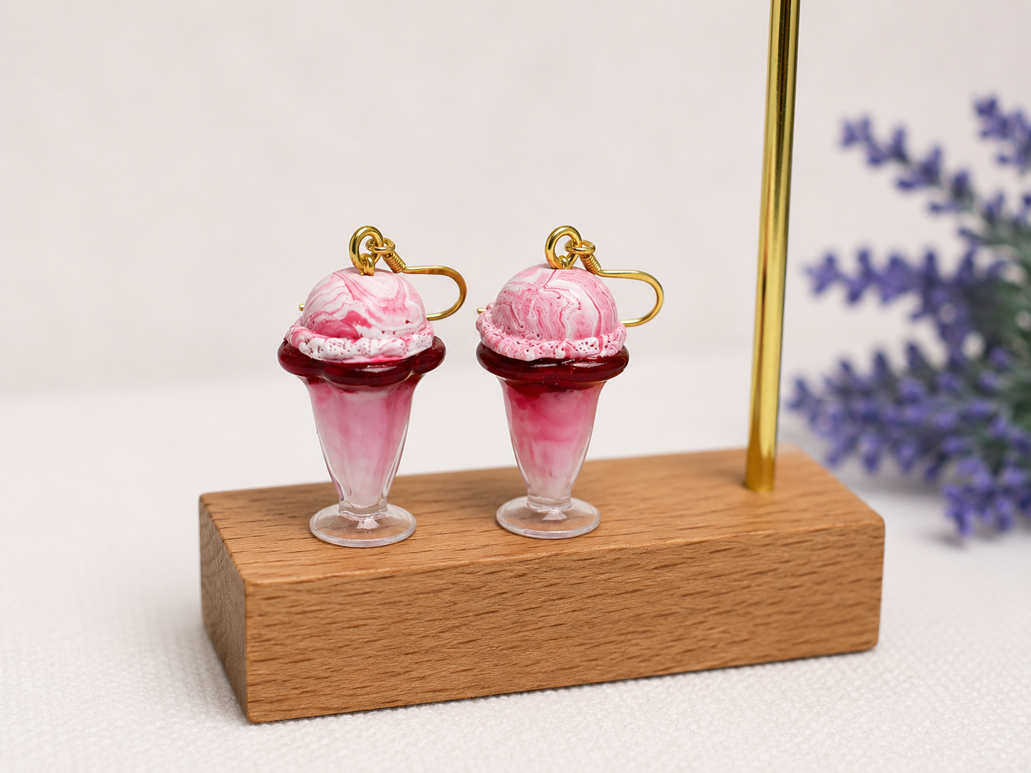 Pair of earrings designed to look like ice cream sundaes, with pink and white marbled scoop on a glass cup, displayed on a wooden block with purple flowers in the background.