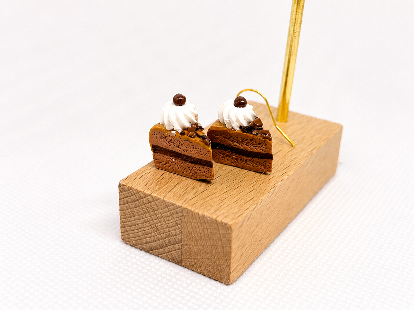 chocolate mousse cake earrings with whipped cream and chocolate shavings on top, placed on a small wooden display stand with a gold decorative rod.