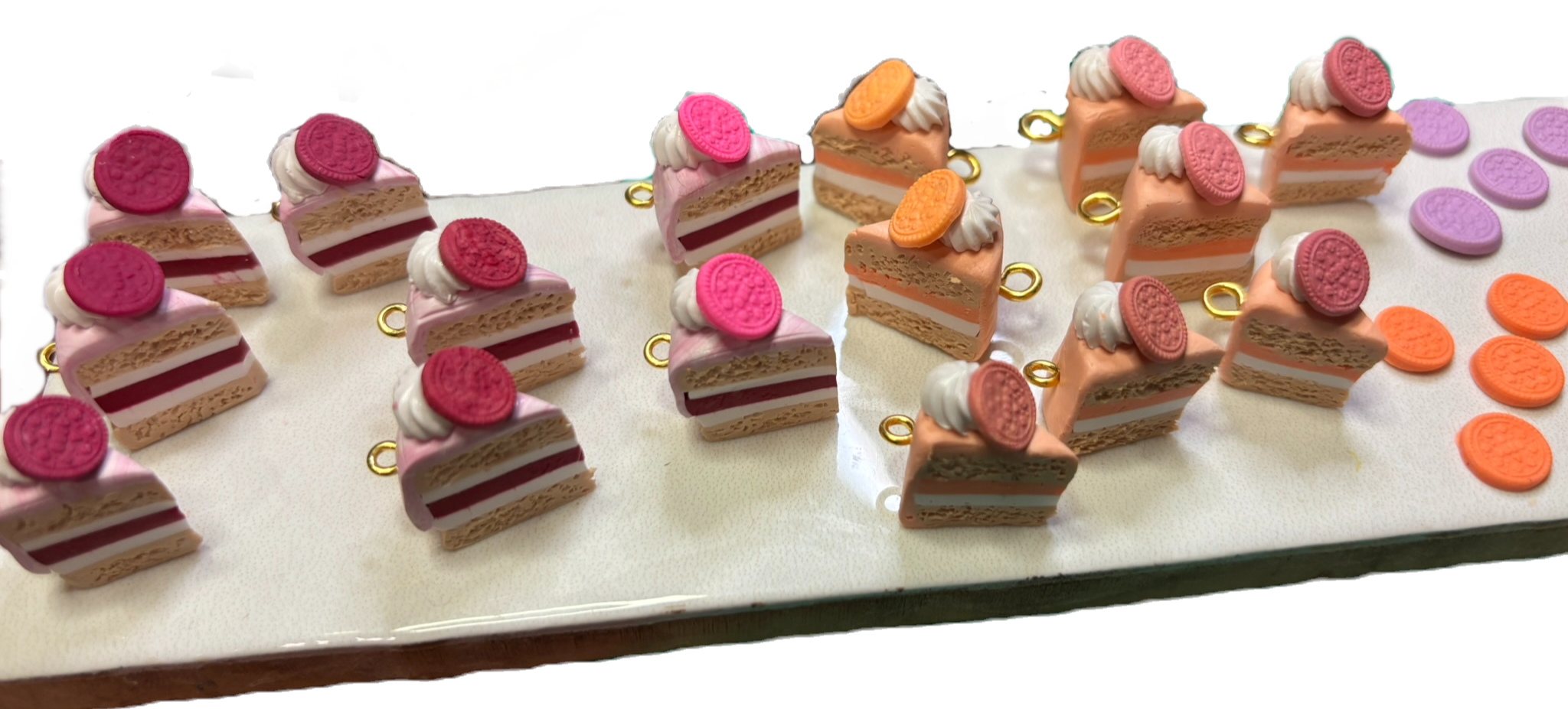 Display of miniature cake and cookie keychains in pink and orange, arranged on a white surface. The cake keychains have layered designs with pink icing and Oreo cookies on top, while the cookie keychains are orange with a cookie on top.