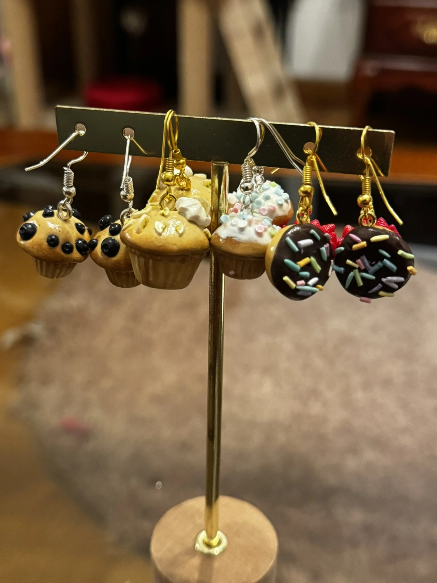 A display stand with five pairs of earrings, featuring cupcake-themed designs, including a cupcake with blueberries, a vanilla cupcake with white frosting and sprinkles, a cupcake with pink and blue decorations, and chocolate cupcakes with colorful sprinkles.