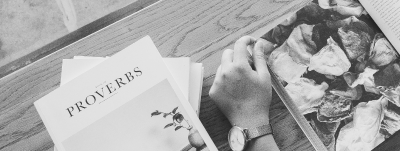 A hand wearing a watch reads a book with a page showing a poem, with other papers on a wooden surface.