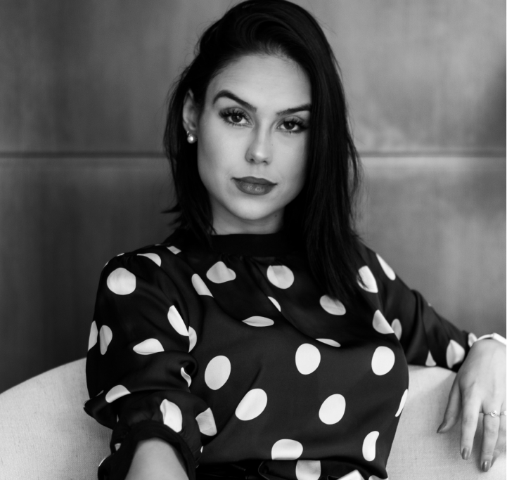 Black-and-white photo of a woman with dark hair wearing a polka dot blouse, seated on a light-colored chair against a wooden background.