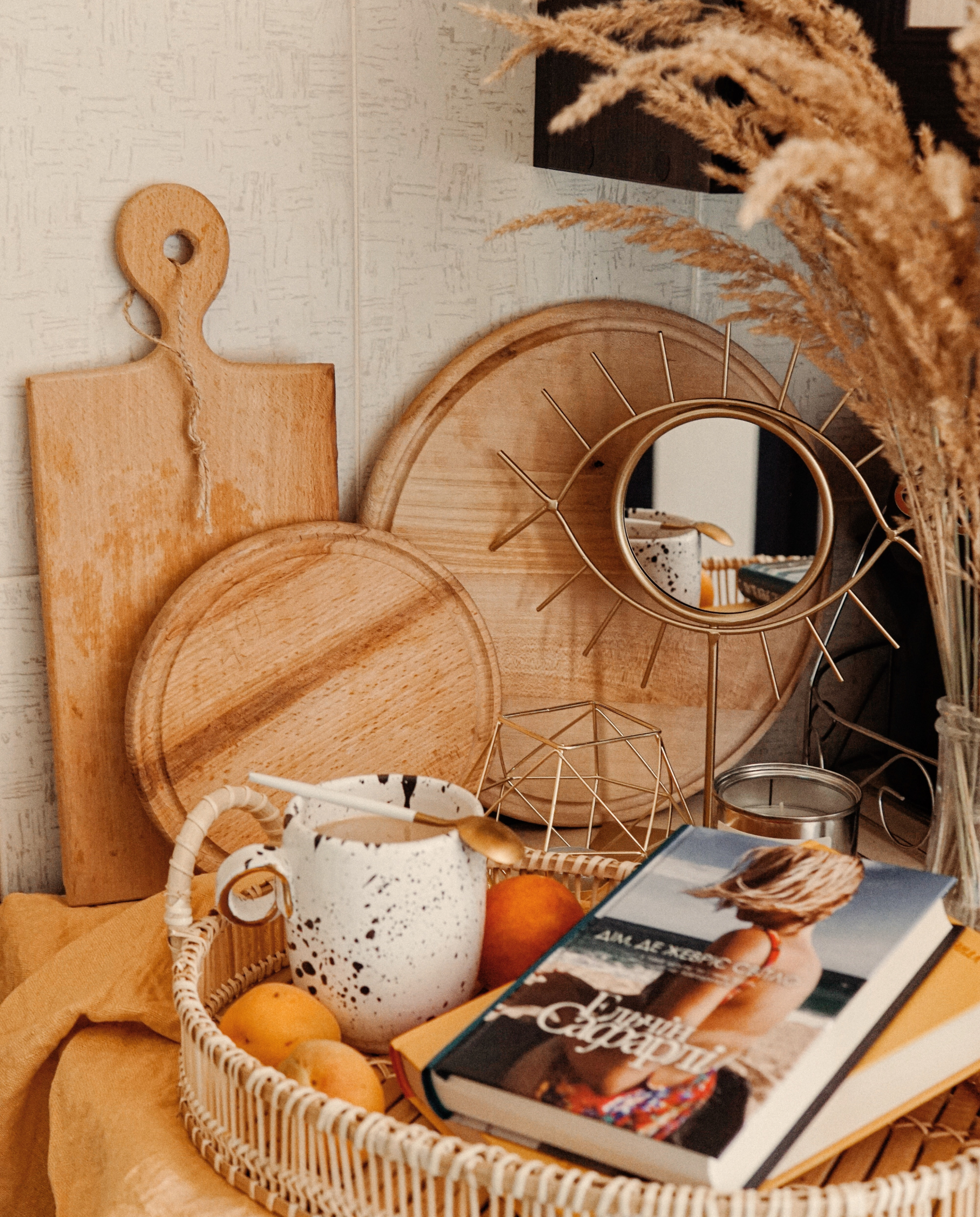 Close-up image of a cozy table setting with wooden cutting boards, a bottle, a white mug with coffee, oranges, a wicker basket, and a book titled 'The Art of...' with a feather on the cover. Dried plants in the background.