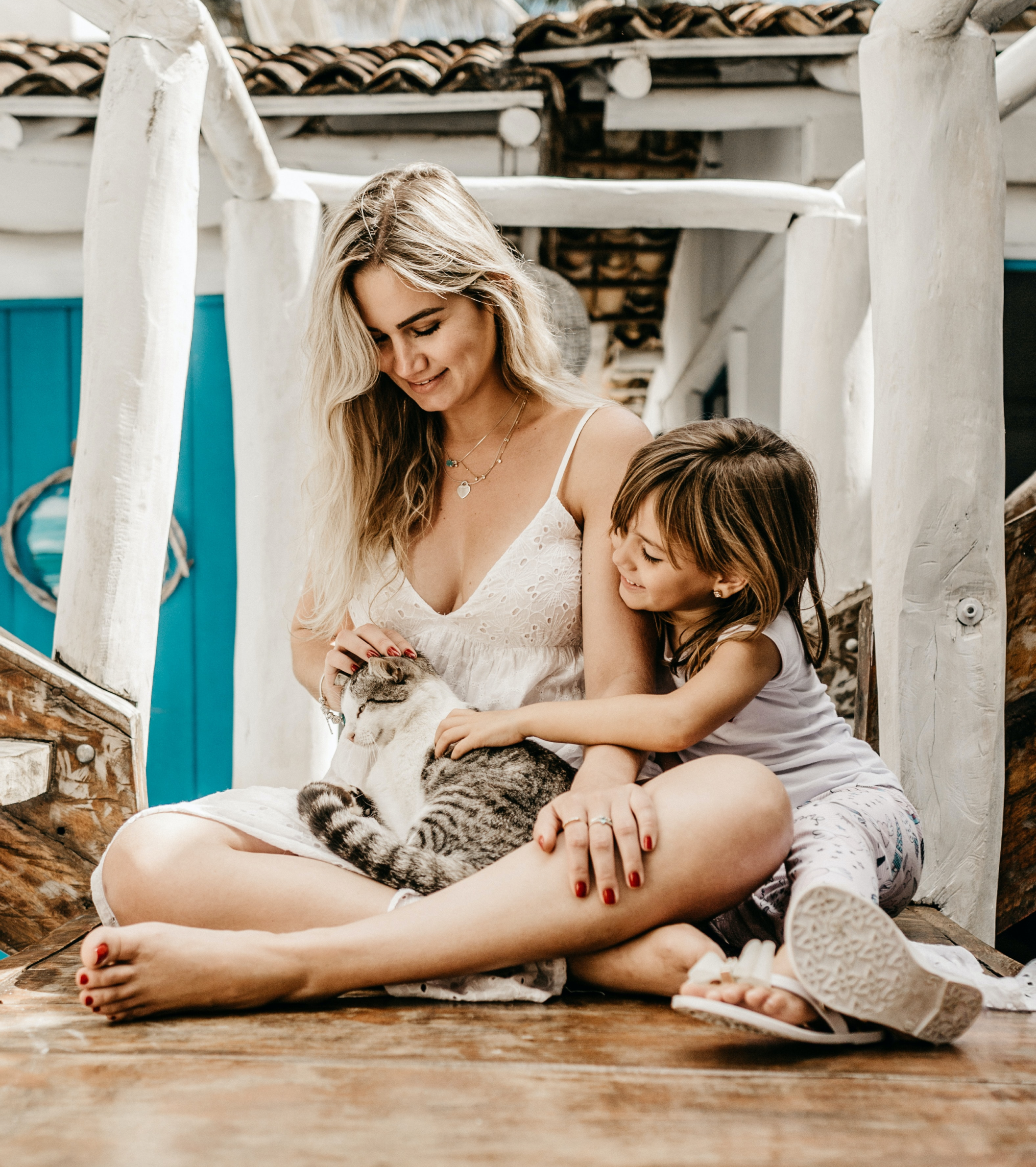 A woman with long blonde hair and a young girl with brown hair sitting together on a wooden floor, petting a black and white cat, with a rustic wooden and white-painted structure in the background.