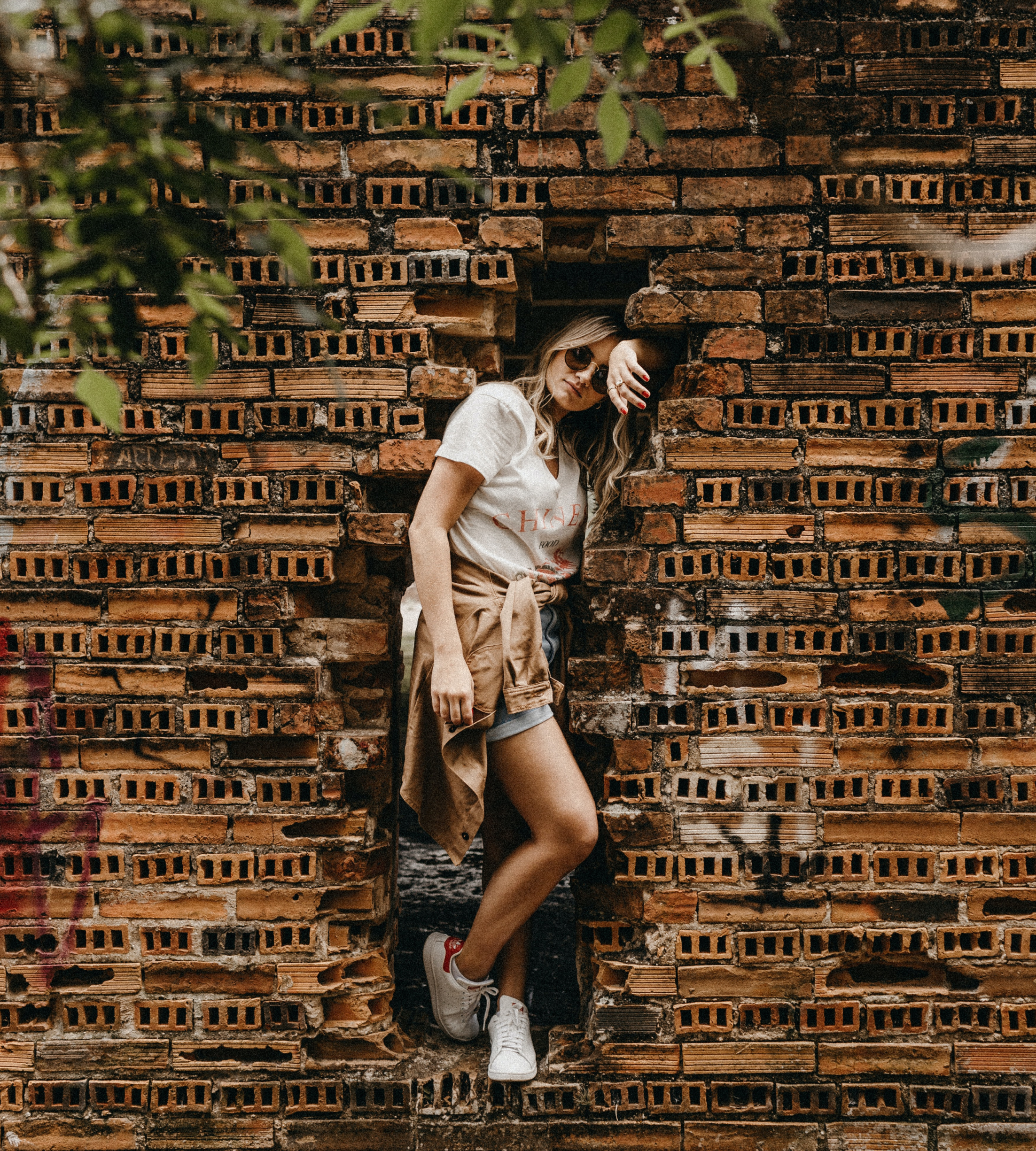 A young woman with long, wavy hair wearing sunglasses, a white t-shirt, shorts, and sneakers, leaning against a rustic brick wall with some greenery in the foreground.
