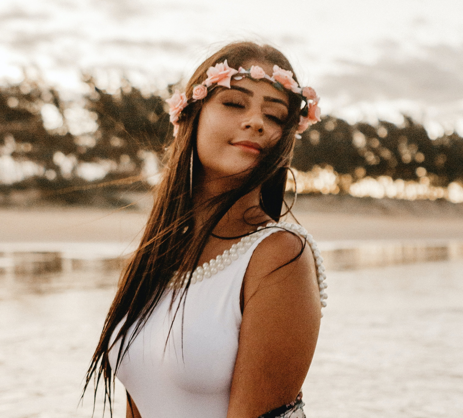 A woman with long dark hair wearing a floral crown and a white dress with pearl accents, standing outdoors during sunset.