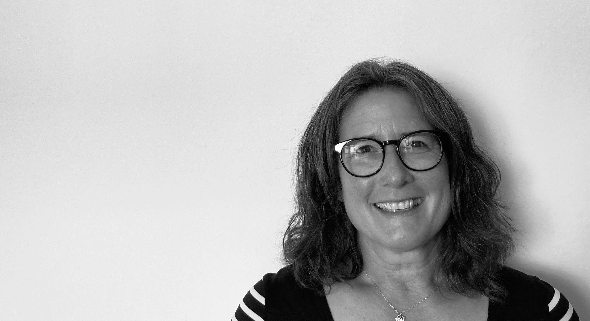Smiling woman with glasses and wavy hair standing in front of a plain wall.