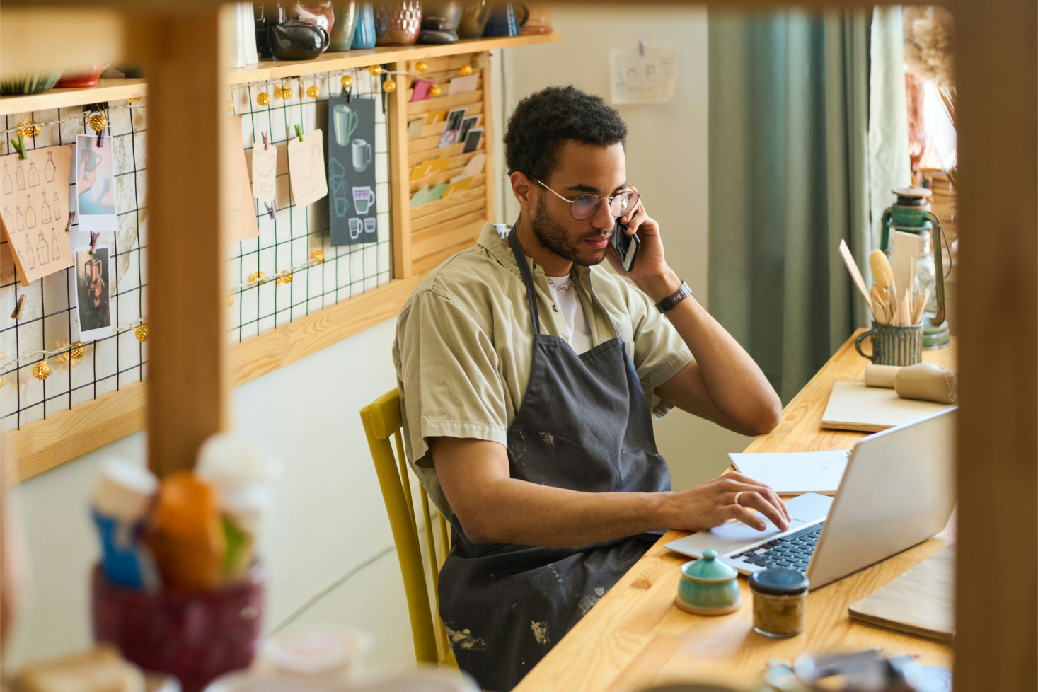 A young man with glasses, wearing an apron, sitting at a wooden table in a cozy, decorated cafe or workspace, talking on the phone, with a laptop open in front of him.