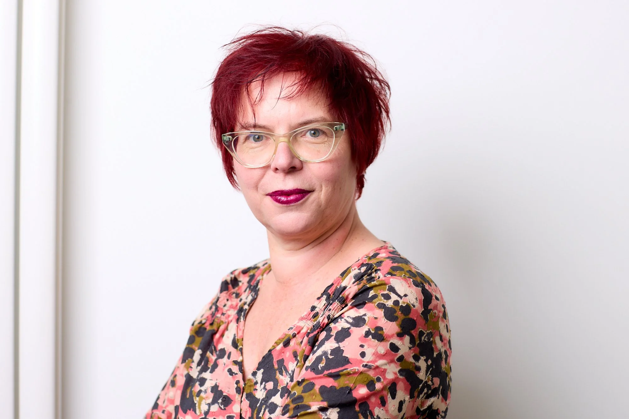 Portrait of a woman with short red hair, wearing glasses and a floral-patterned top, standing against a plain white wall.