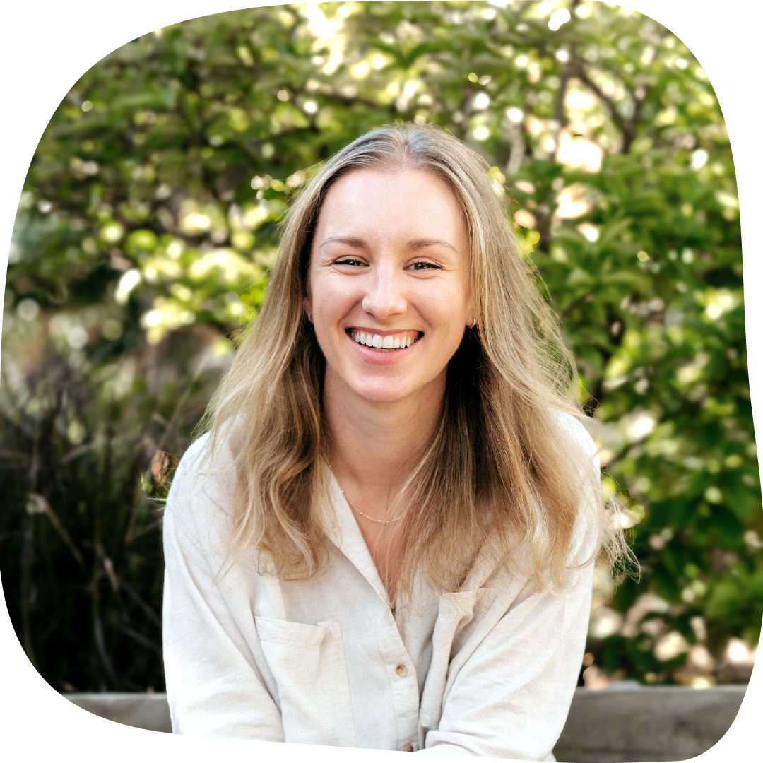 Lydia with long blonde hair, smiling and wearing a beige shirt, outdoors with green foliage in the background.