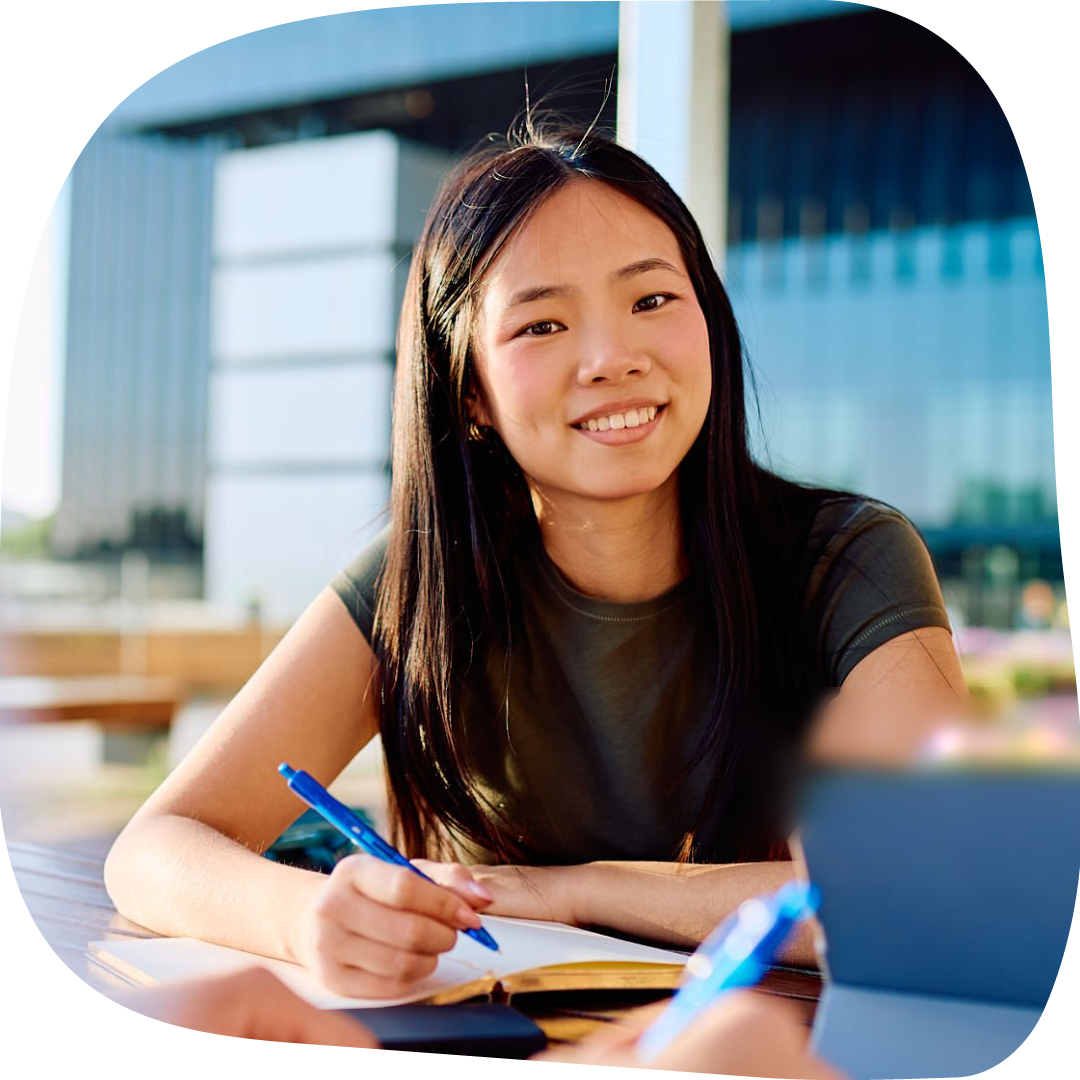 Young female student smiling while writing in a notebook outdoors with a modern school in the background.