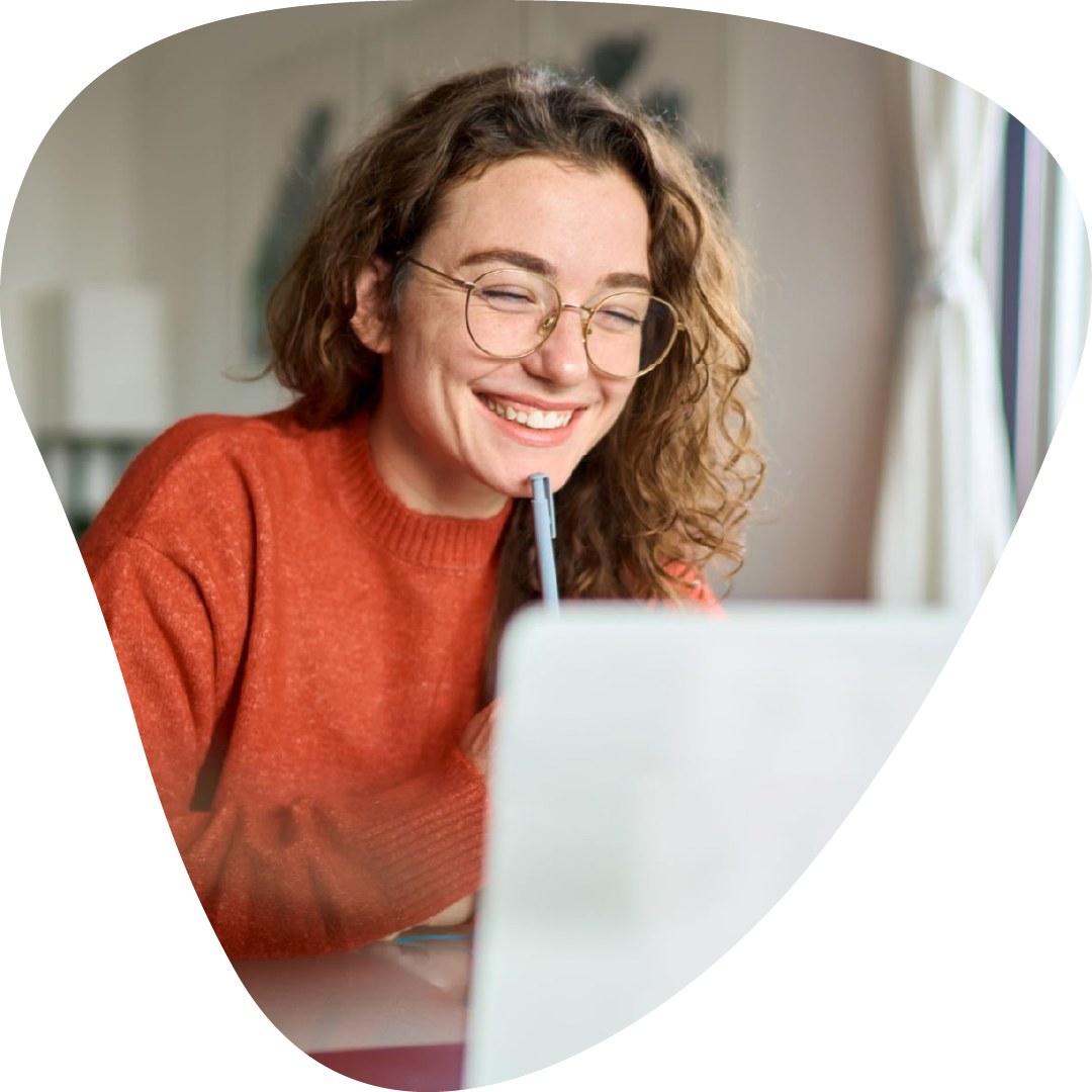 A female student with curly brown hair, wearing glasses and an orange sweater, smiling while learning Spanish on a laptop screen in a cozy room.
