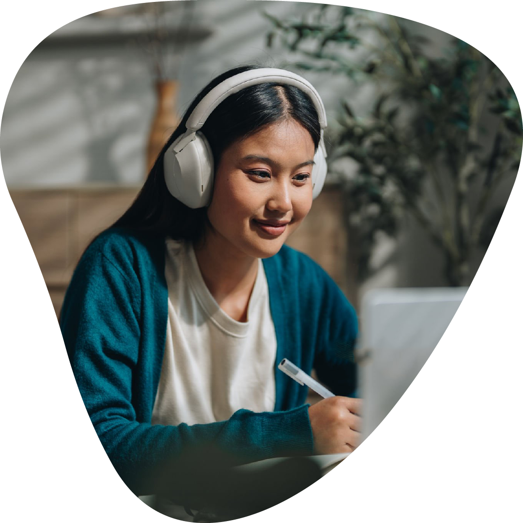 A young female student with long dark hair, wearing a teal cardigan and white shirt, being tutored with white headphones, sitting at a table with a laptop and taking notes.