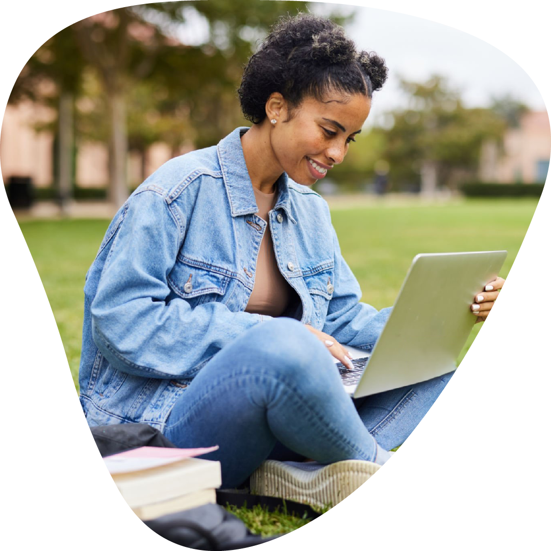 A young woman with short, curly hair sitting on the grass in a park, smiling while working on a silver laptop. She is wearing a light blue denim jacket and matching jeans, with books and a backpack nearby.
