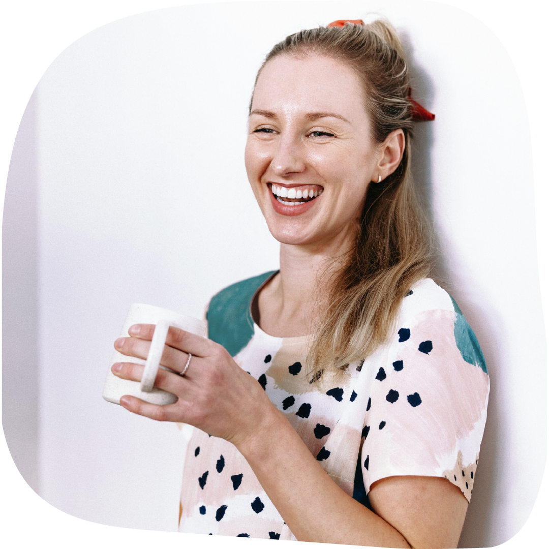 Lydia with light brown hair, smiling and holding a white mug, standing against a white wall, wearing a colorful polka-dot shirt.