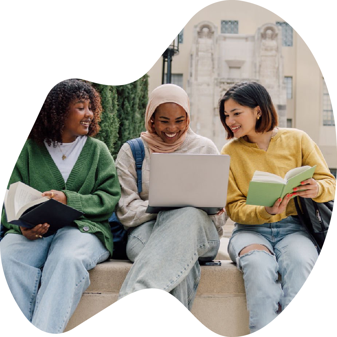 Three young women student sitting on a bench outside of school, smiling and looking at a laptop, with books in their hands, in front of a historic school building.