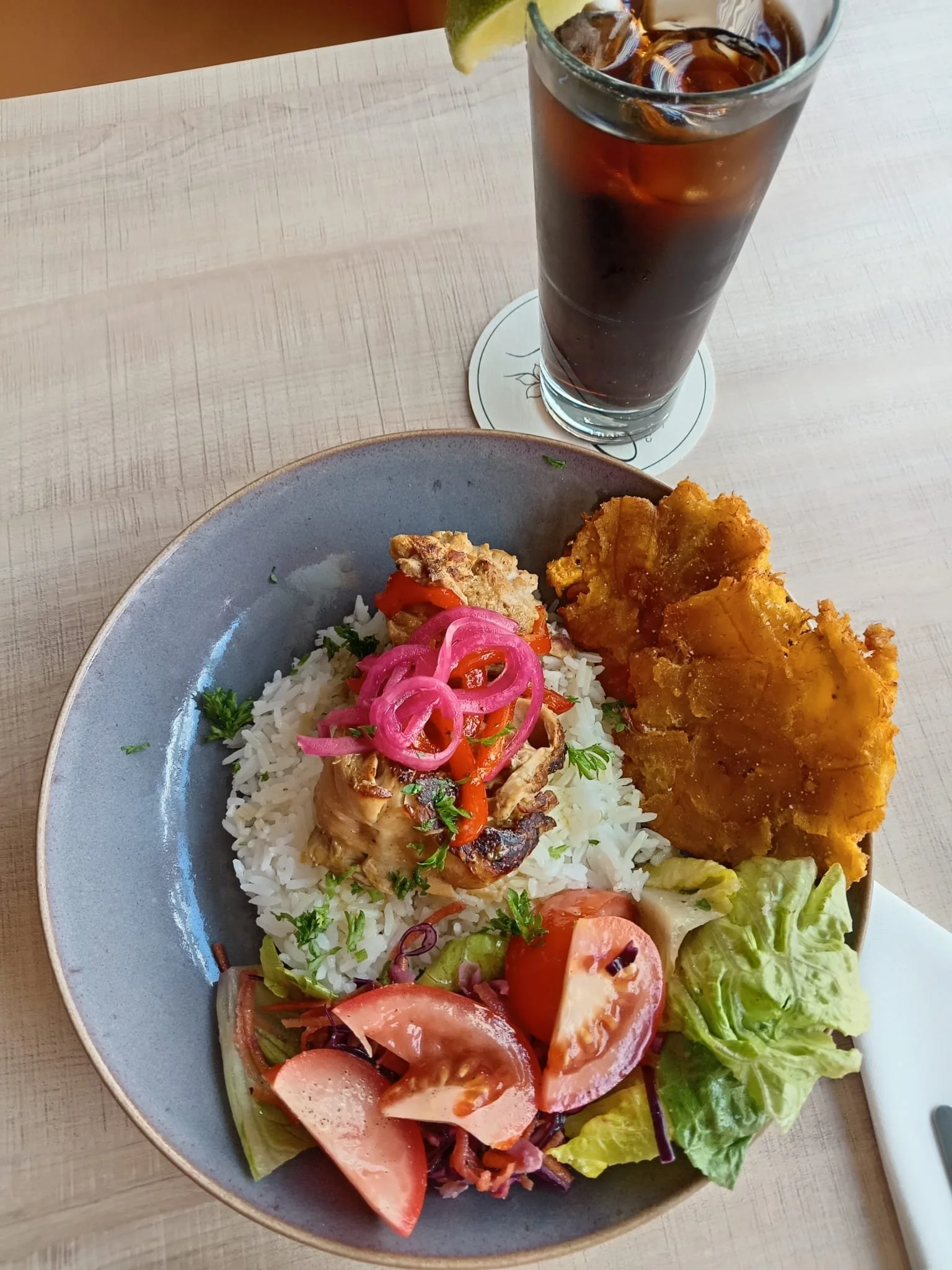 A plate of rice with grilled chicken topped with pickled red onions and red pepper slices, accompanied by a salad with lettuce, tomato, and cucumber slices, and a glass of dark-colored soda with ice and lime wedge.