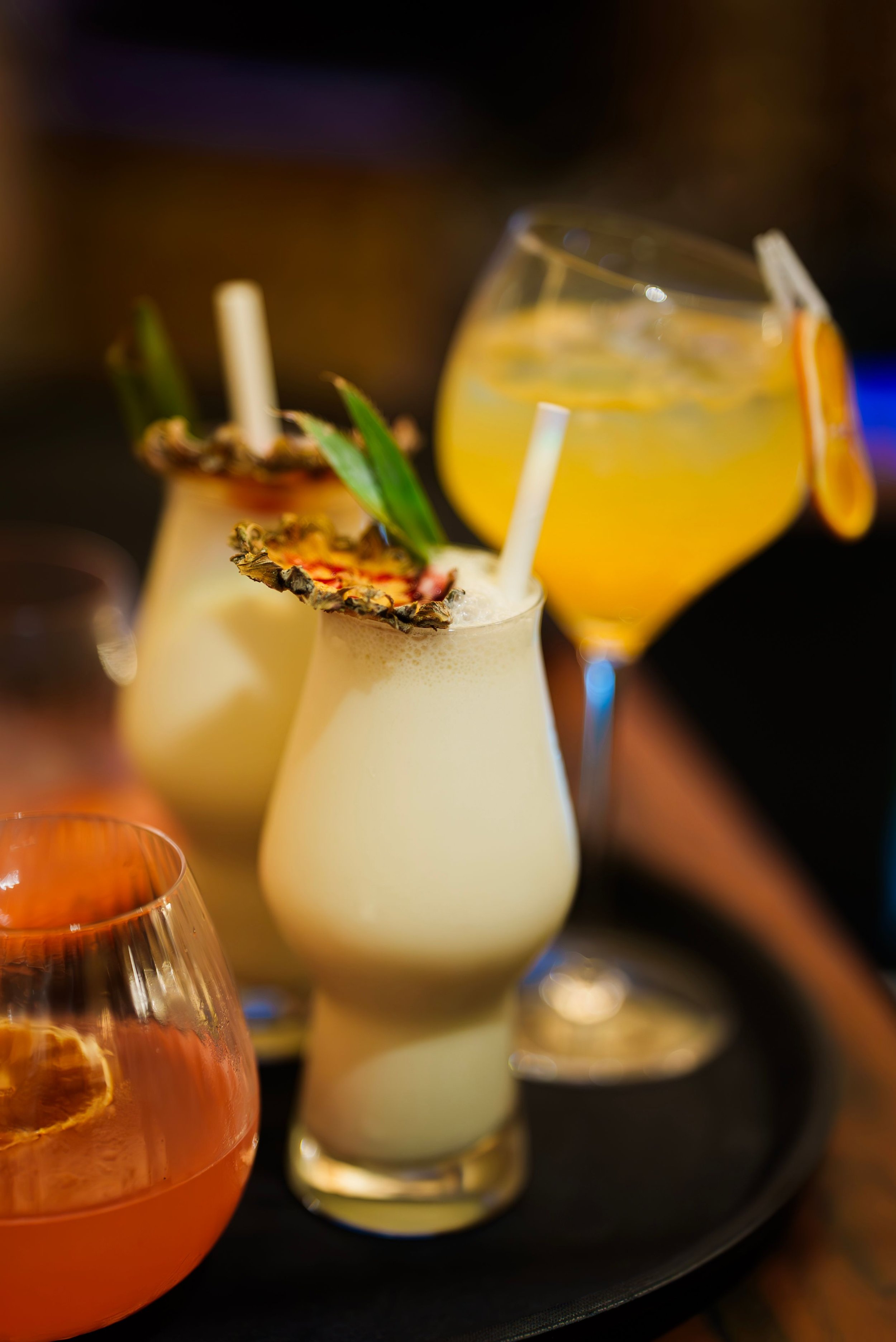 Three tropical cocktails with garnishes on a black tray. Two have pineapple slices and green leaves, one features a slice of orange. All drinks have straws.