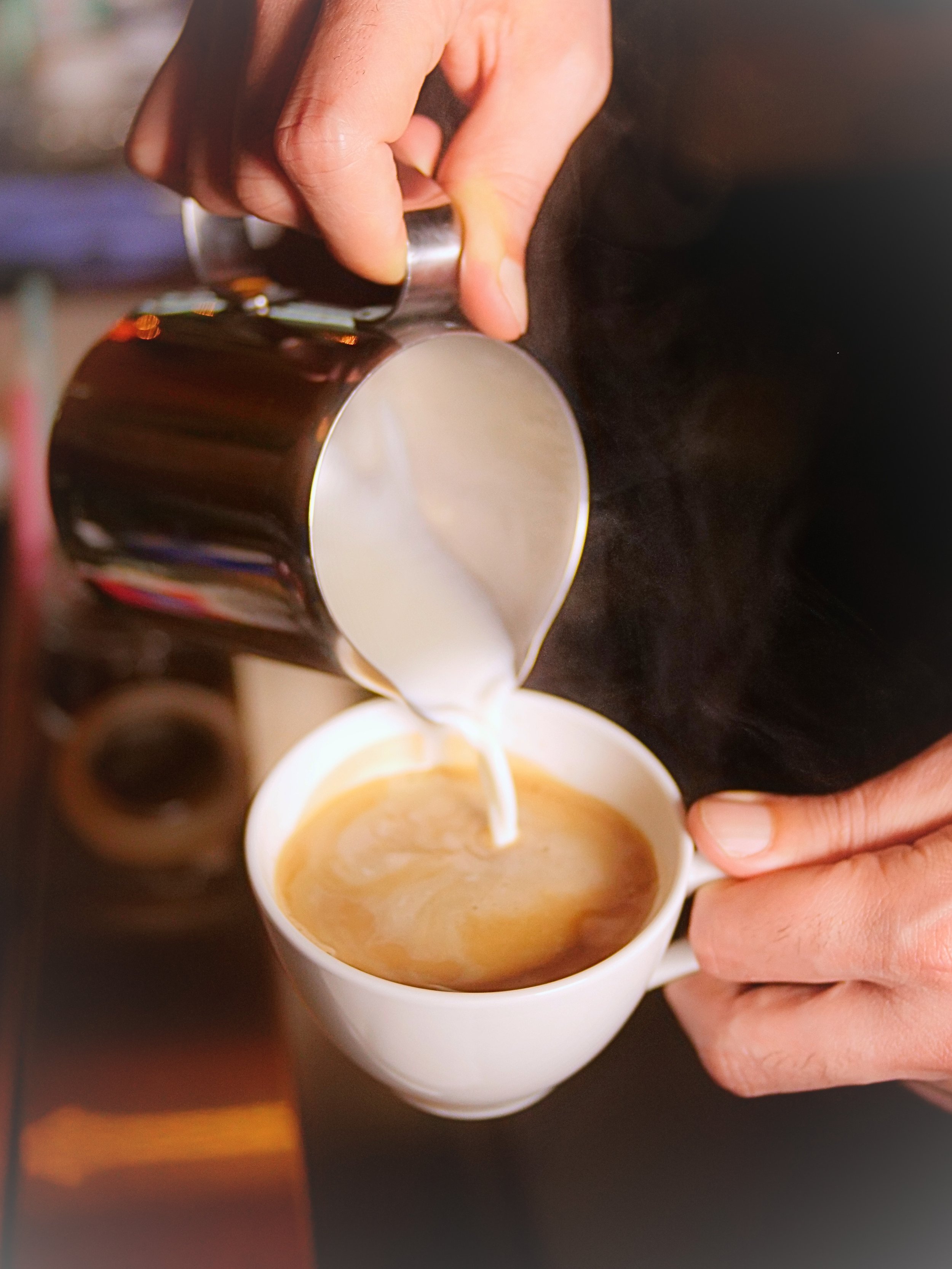 Close-up of a person pouring a hot beverage from a metallic pitcher into a white mug, which is being held by the person's hand. The beverage appears to be coffee or espresso.