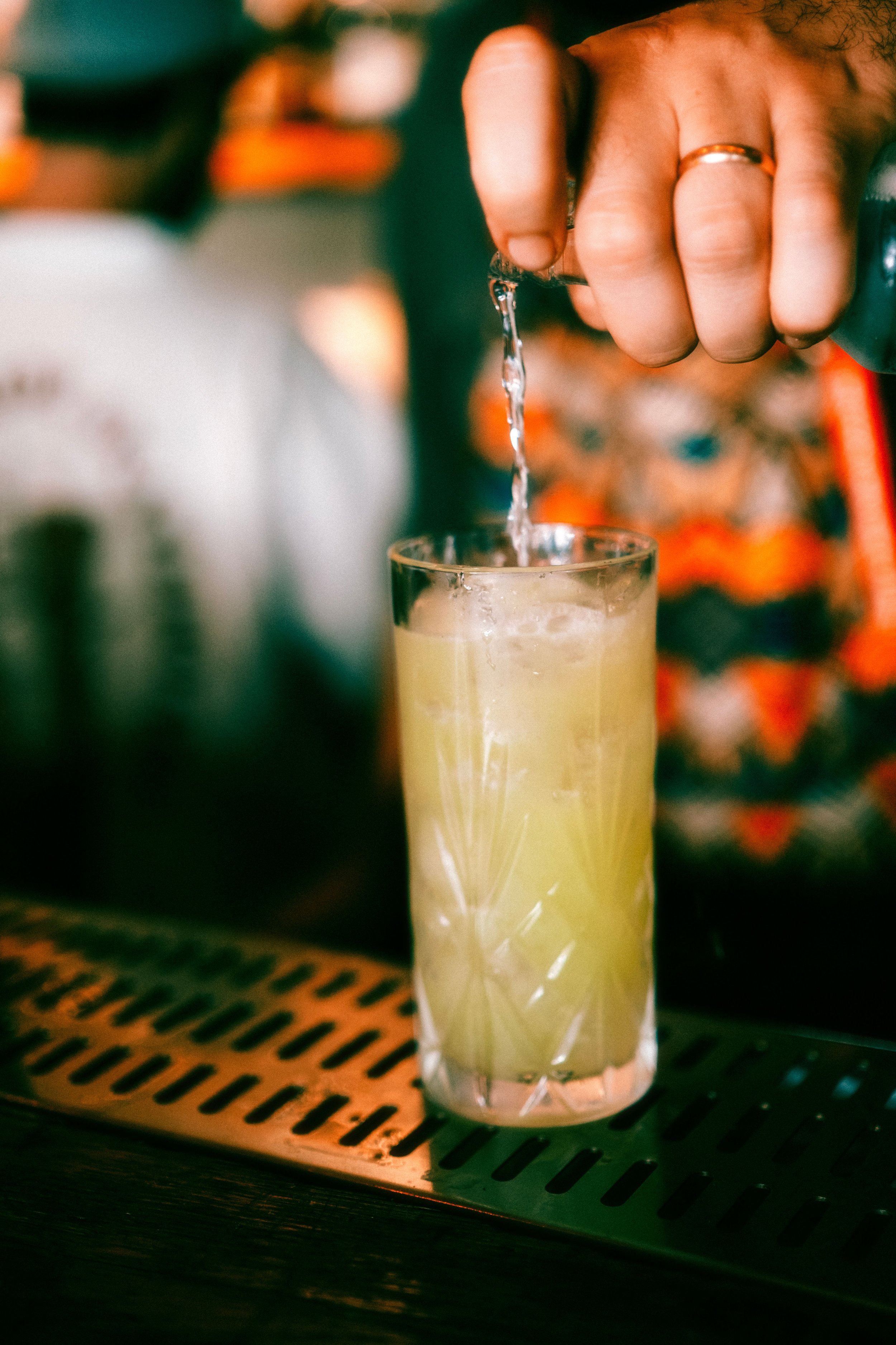 Close-up of a hand pouring a yellow-colored drink into a tall glass.