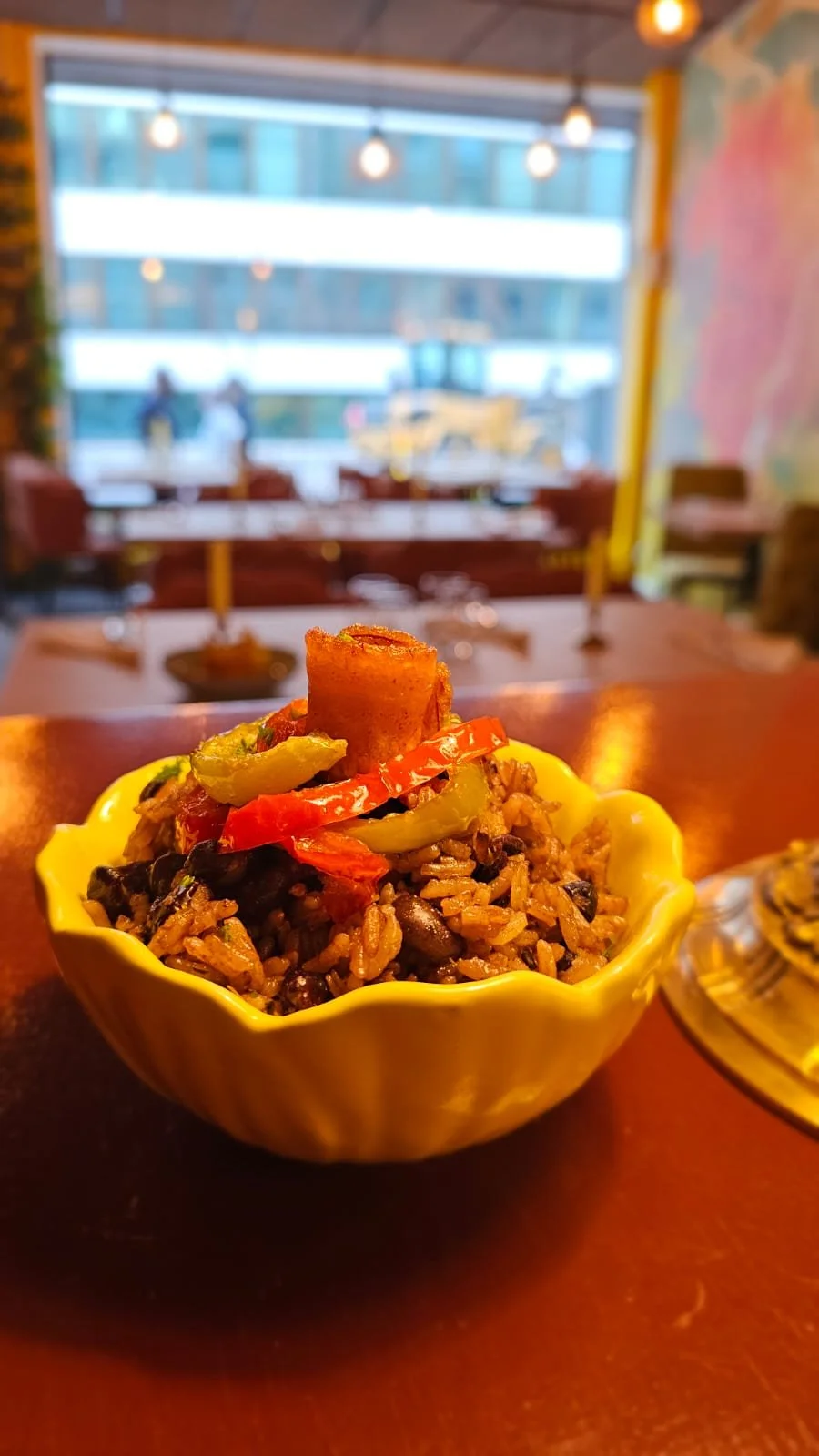 Bowl of black bean and rice salad topped with chopped vegetables and a fried plantain slice, on a dining table in a restaurant with large window and modern decor.