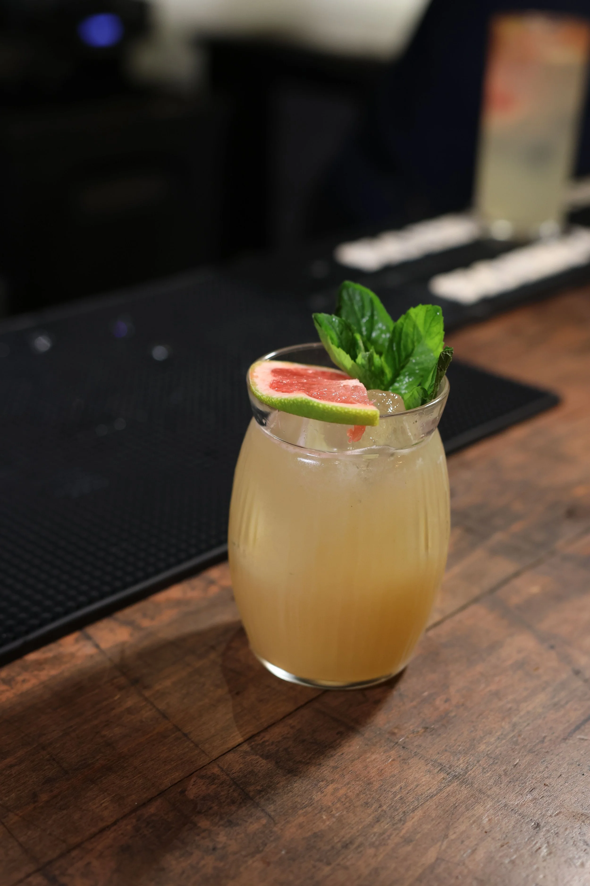 A cocktail in a clear glass with a lime wedge and a sprig of mint garnishing the drink, set on a wooden surface.