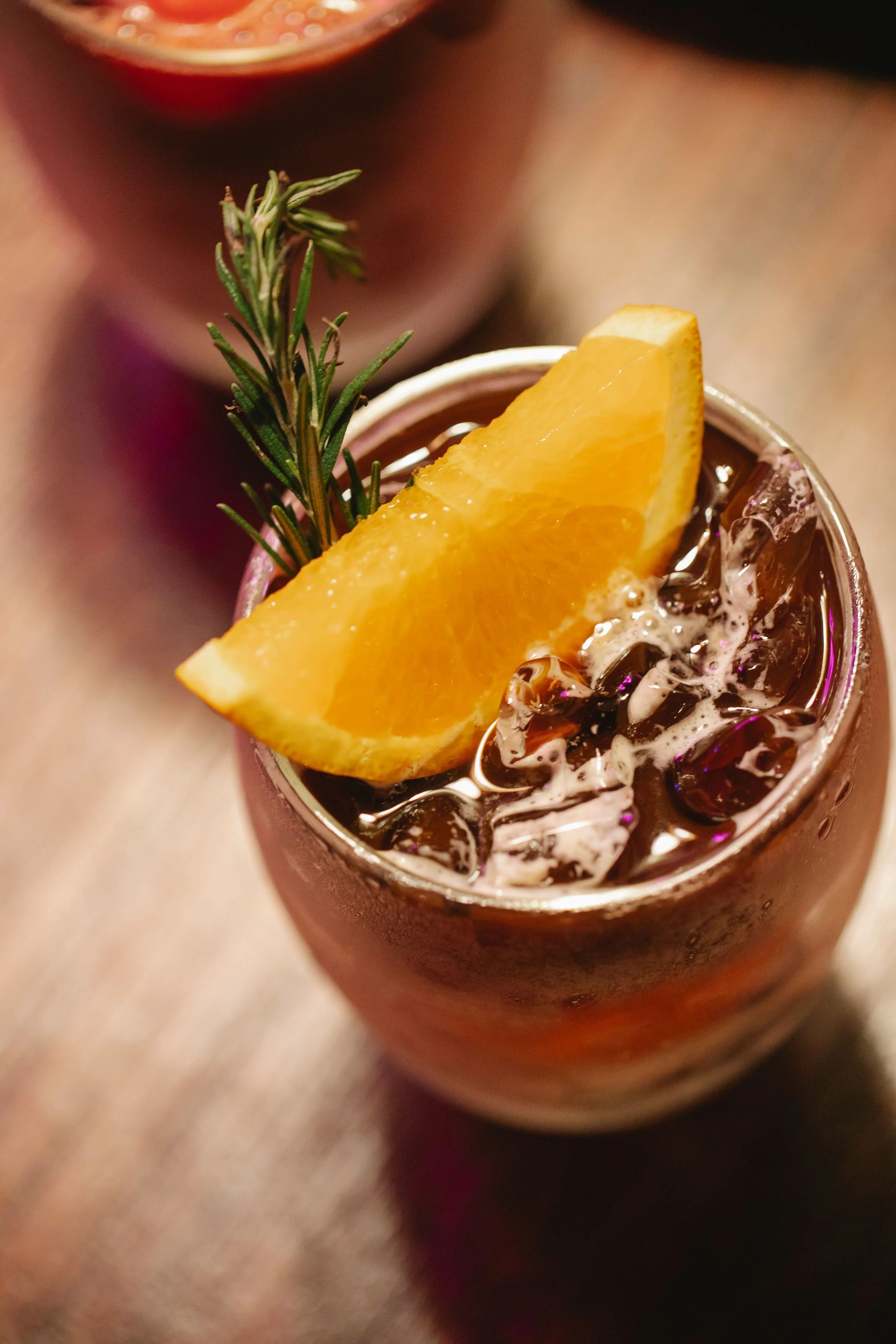 Close-up of a cocktail garnished with an orange wedge and a sprig of rosemary, with an ice-filled glass and a blurred background.