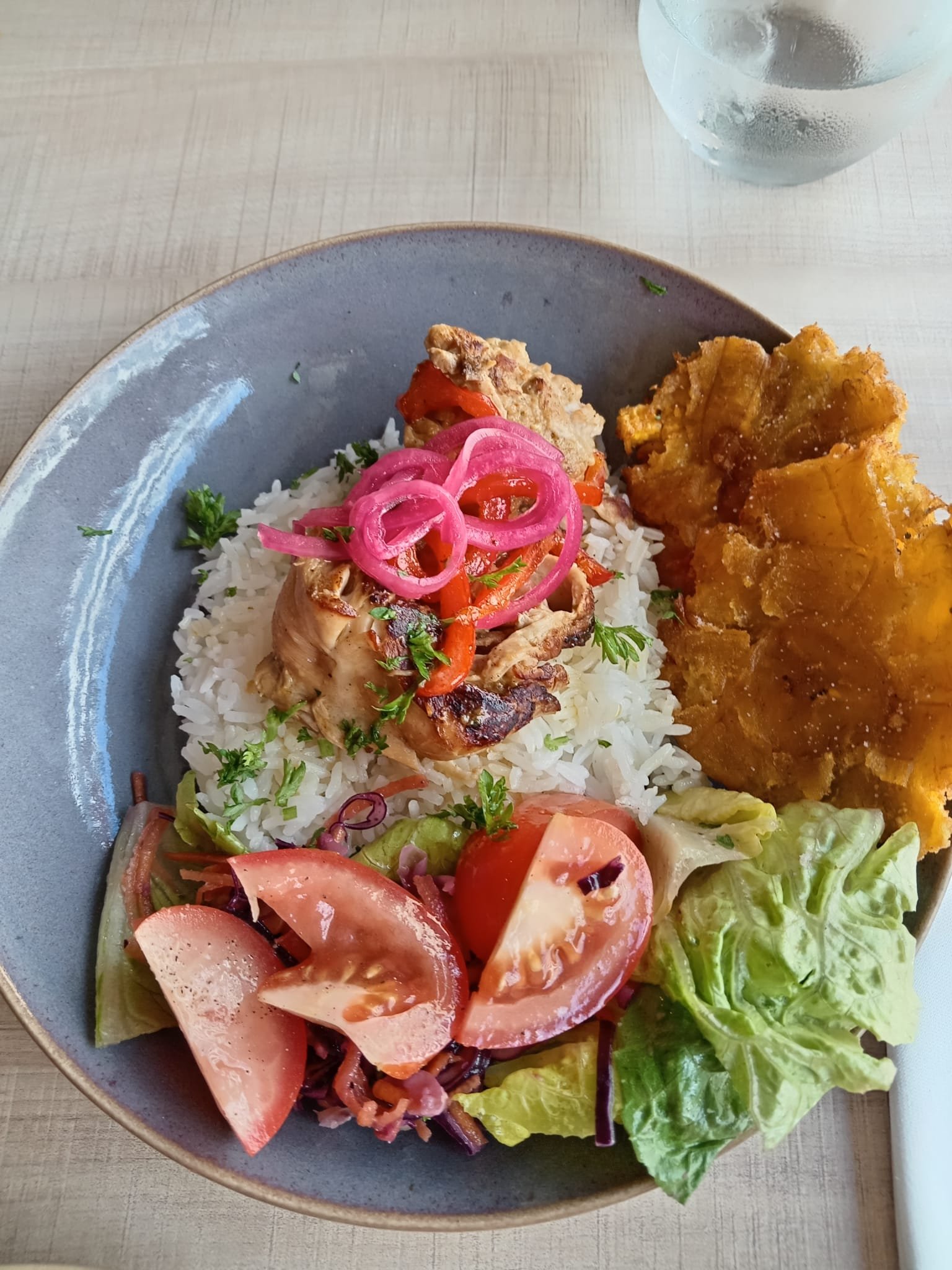 A plate with rice, grilled chicken topped with pickled red onions, a fried plantain, and a fresh vegetable salad with tomatoes, lettuce, and radish slices.