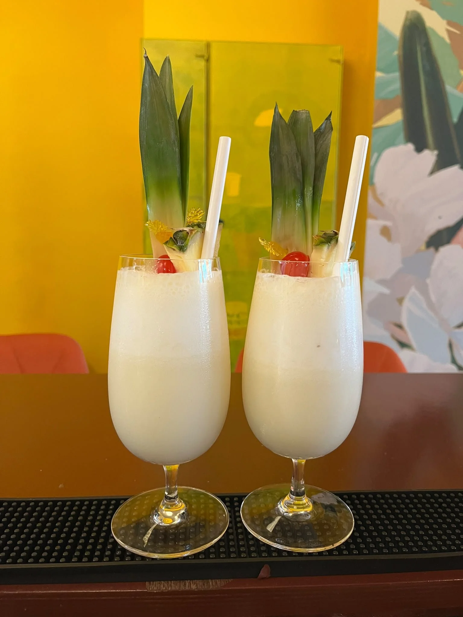 Two tropical drinks garnished with pineapple, cherry, and straw, displayed on a black textured surface against a colorful background.