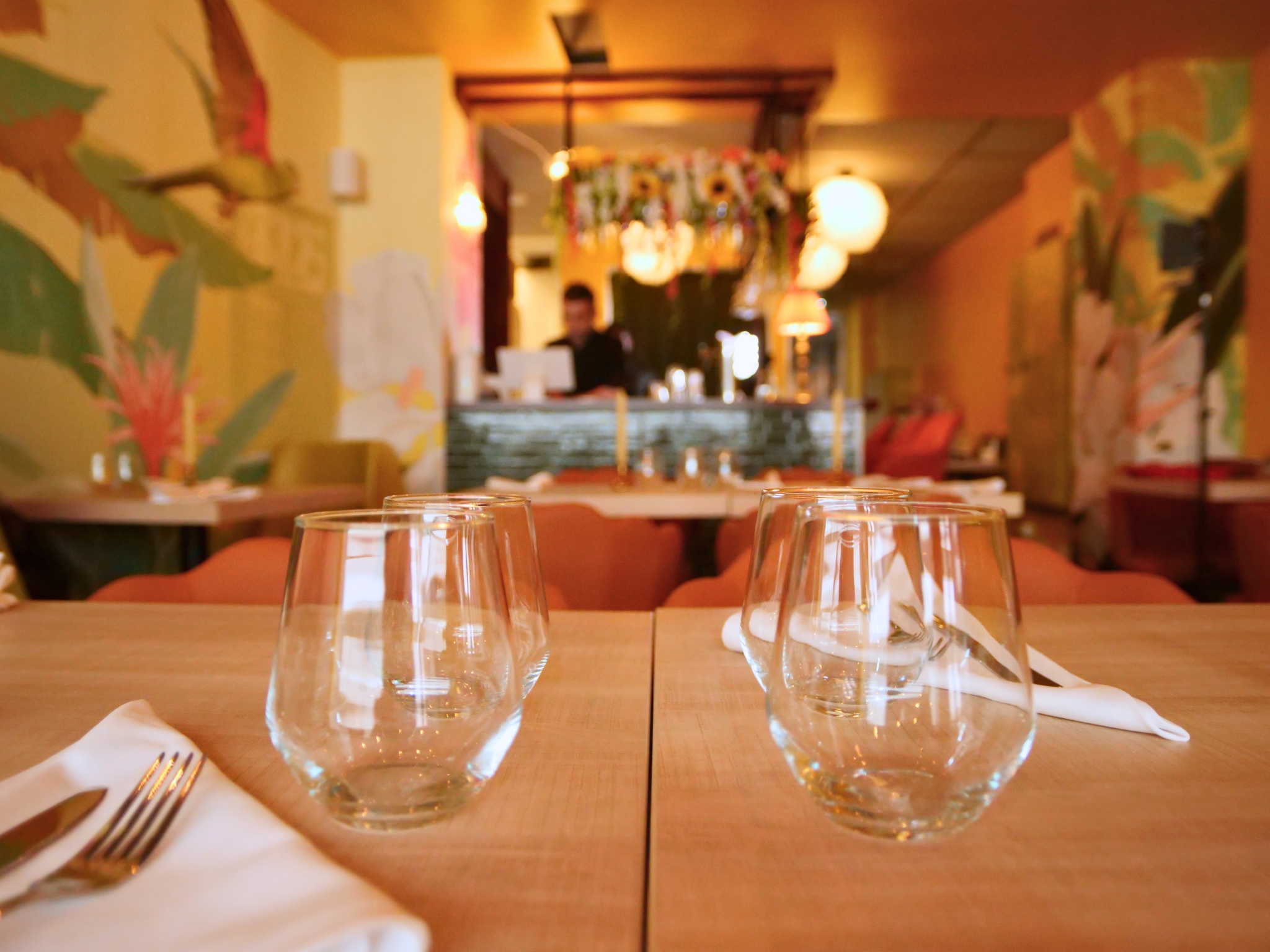 The image shows a restaurant or dining area setting with a wooden table, empty glasses, a fork and a napkin in the foreground. In the background, there is a reception or bar counter with a person standing behind it, and hanging light fixtures and dec