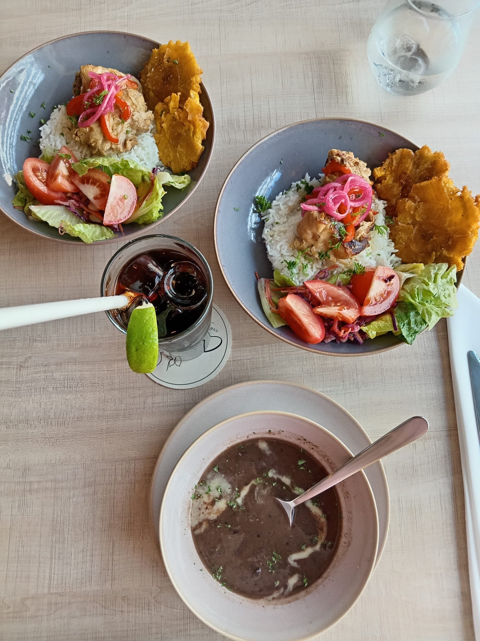 Two bowls of rice with chicken, fried plantains, and salad with sliced tomatoes and lettuce. One bowl has pink pickled onions. A glass of dark soda with a lime wedge, a glass of water, and a bowl of dark soup with herbs are on the table.