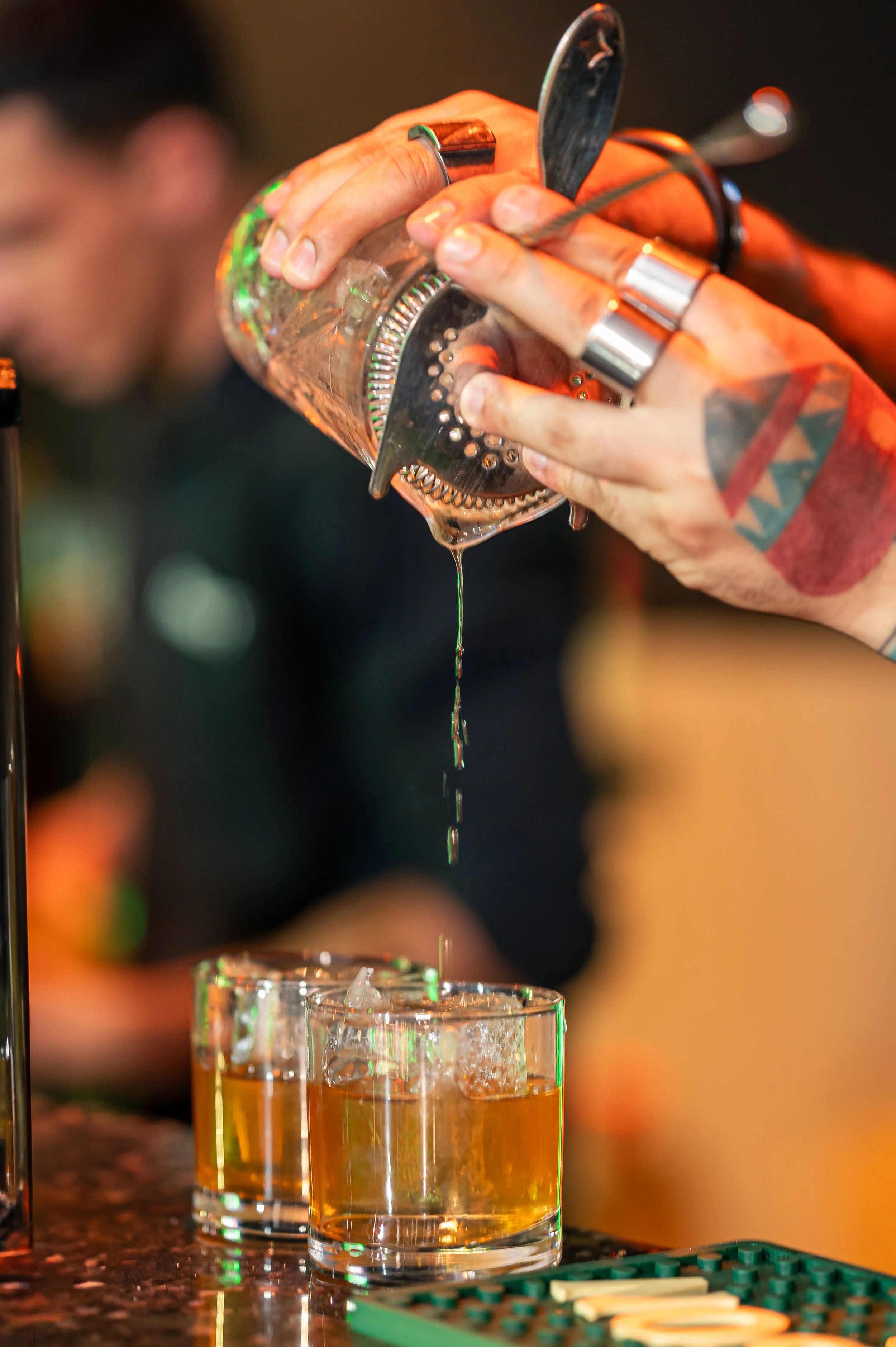 A person is pouring a shot of amber-colored liquor from a stainless steel cocktail shaker into a glass with ice. The person's hands are decorated with rings and rings tattoos, and they are using a bar spoon to help pour.