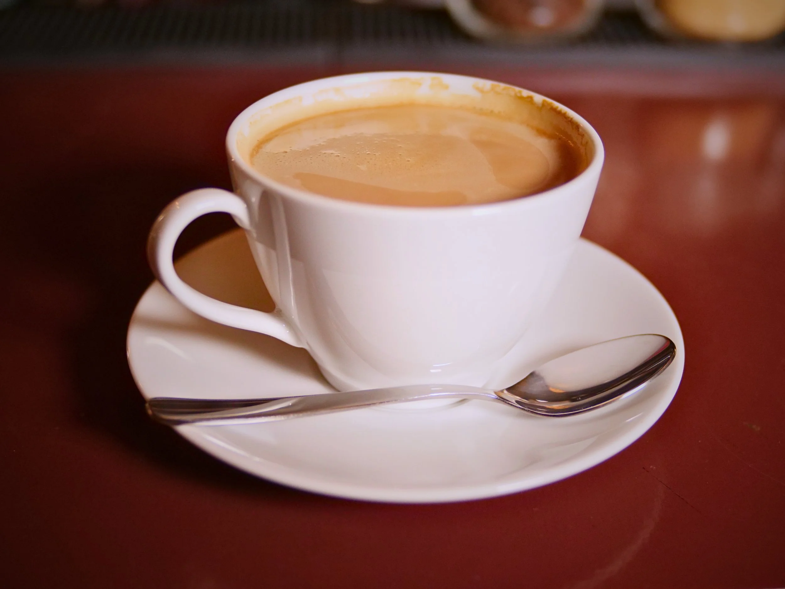 A white ceramic cup full of coffee with foam on top, placed on a matching saucer with a spoon on it, on a dark wooden table.