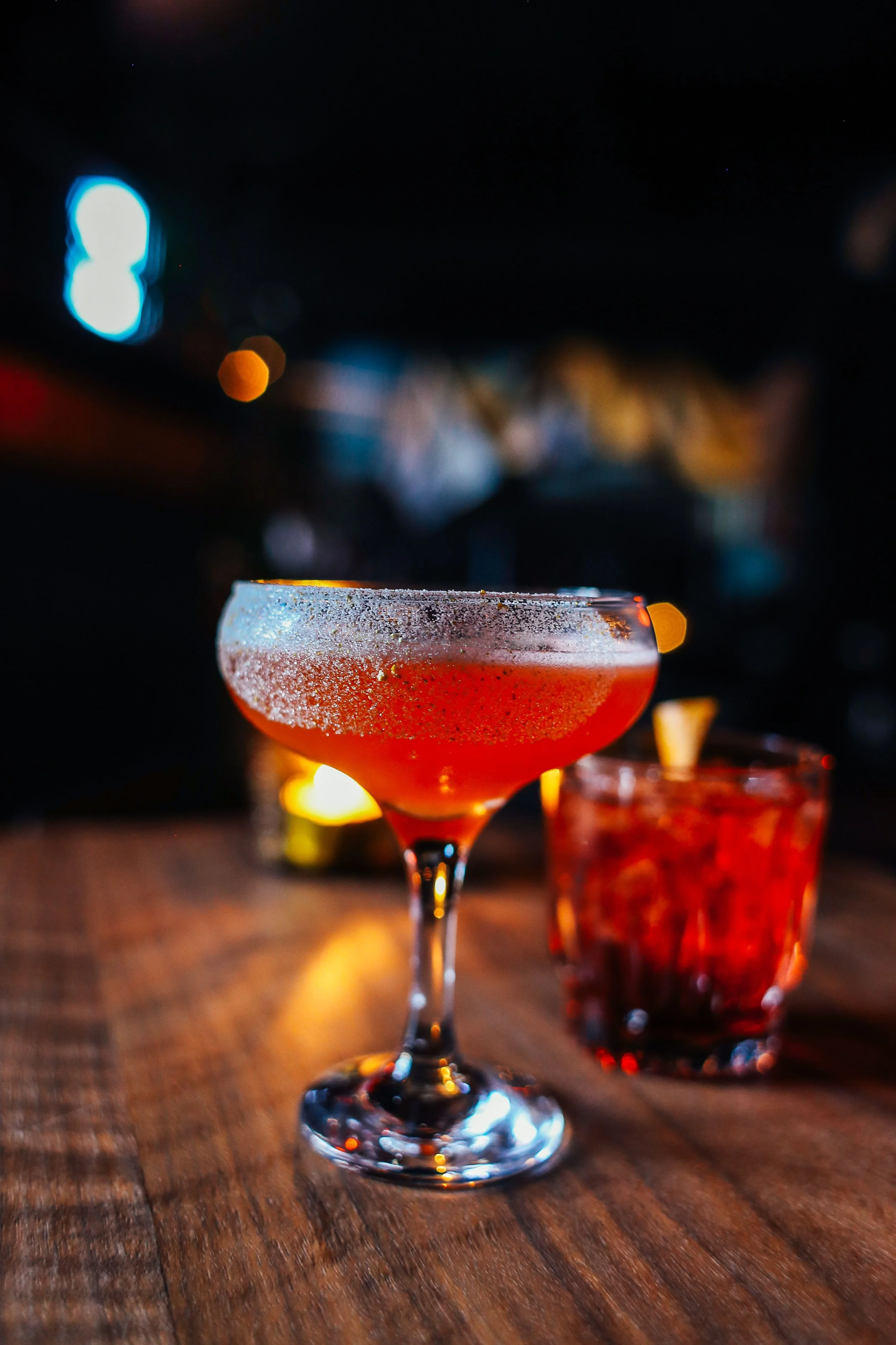 A pink cocktail with a salted rim in a coupe glass on a wooden table at night, with blurred background lights.