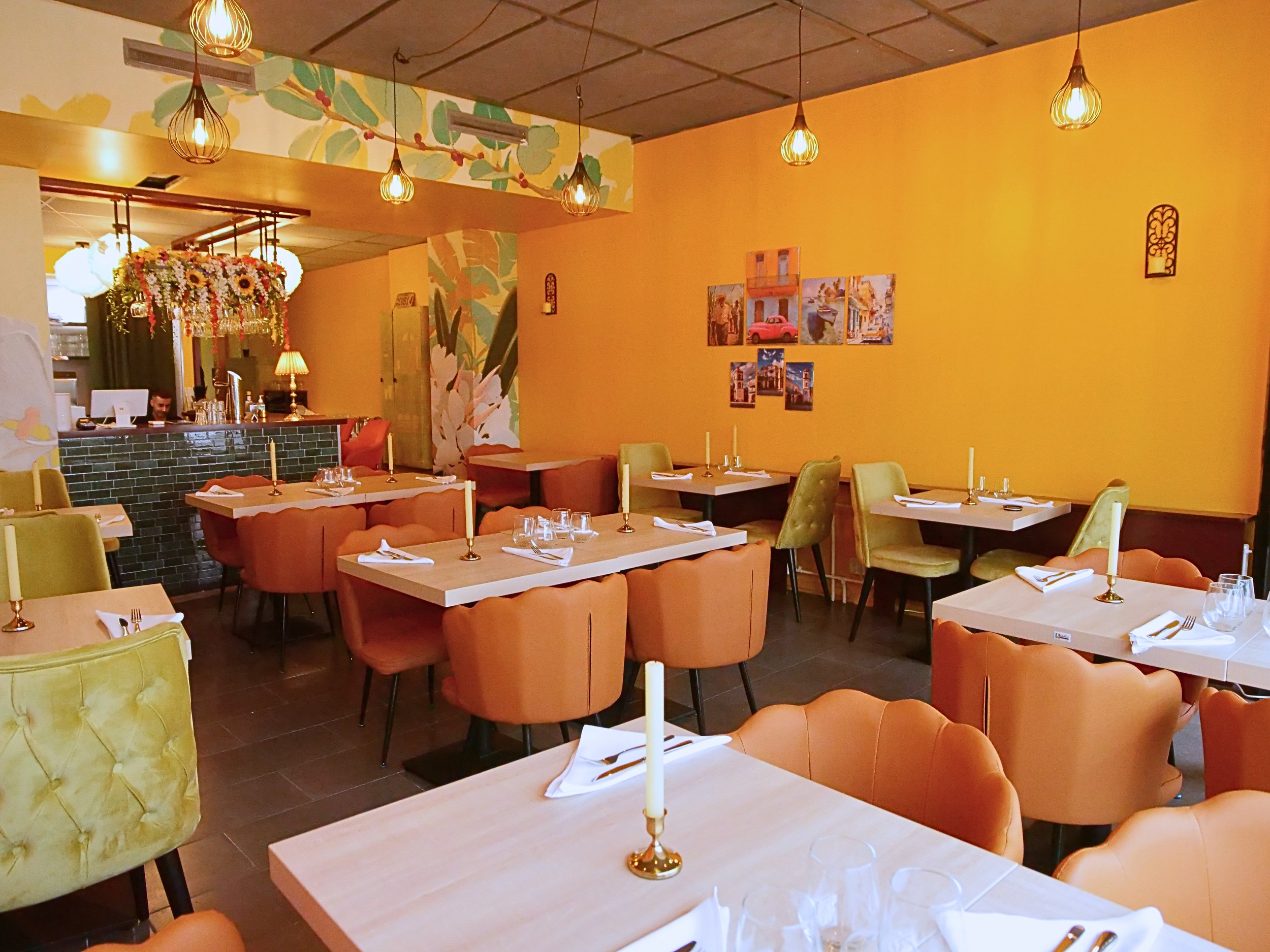 Interior of a restaurant with yellow walls, decorated with paintings and wall art. Several tables are set with white napkins, cutlery, and candlesticks. Various chairs, some in green and others in a peach hue, are arranged around the tables. Hanging 