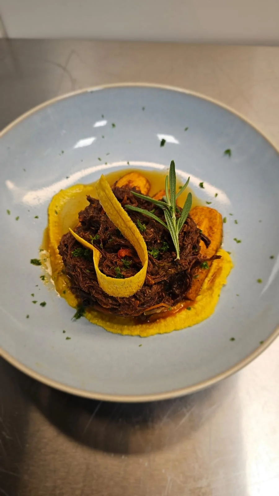 A plated dish of braised beef with rosemary garnish, served over mashed potatoes and mashed yellow vegetables, garnished with herbs and lemon peel.