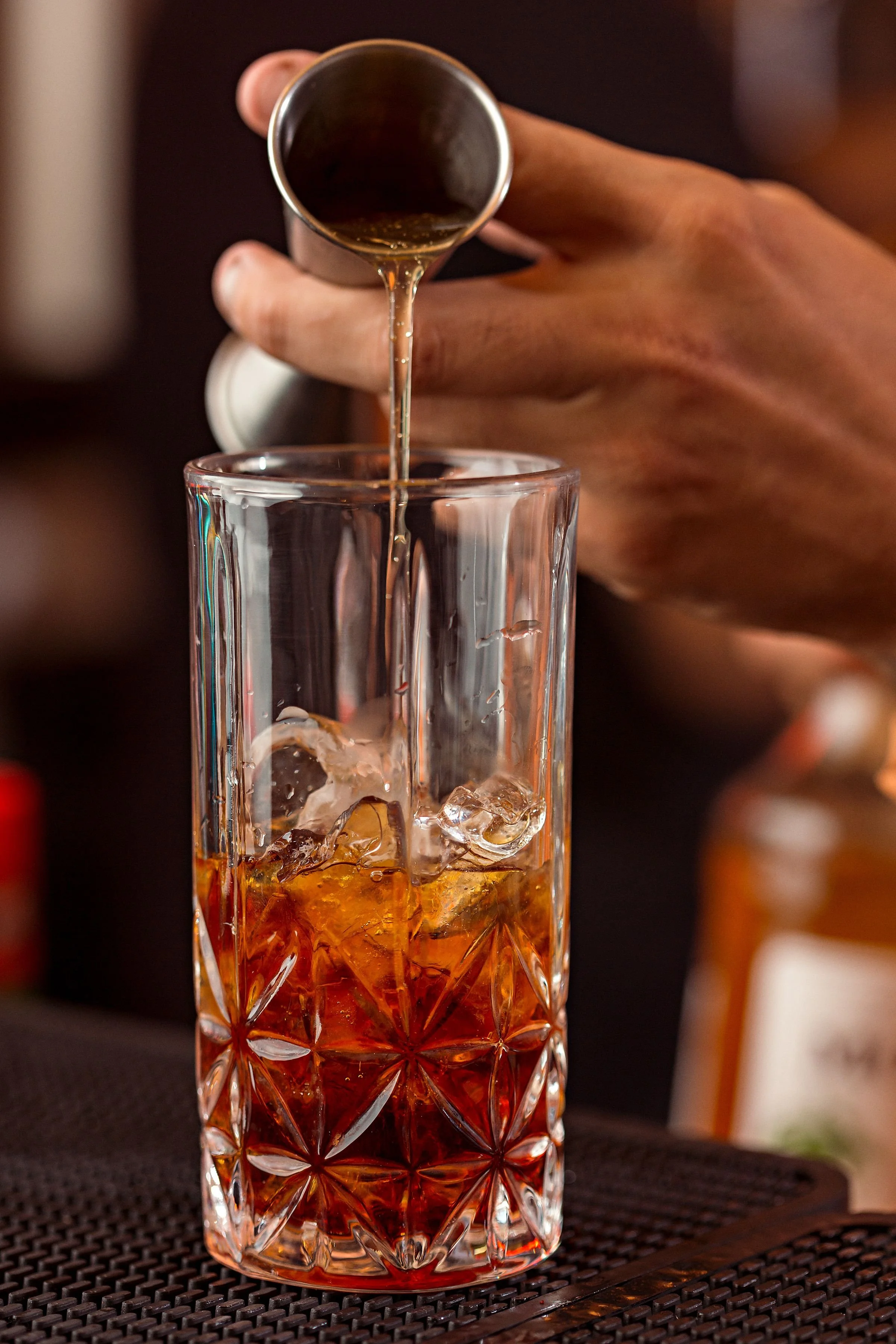 A hand pouring a dark beverage from a small metal pitcher into a tall, patterned glass filled with ice, on a dark textured surface.
