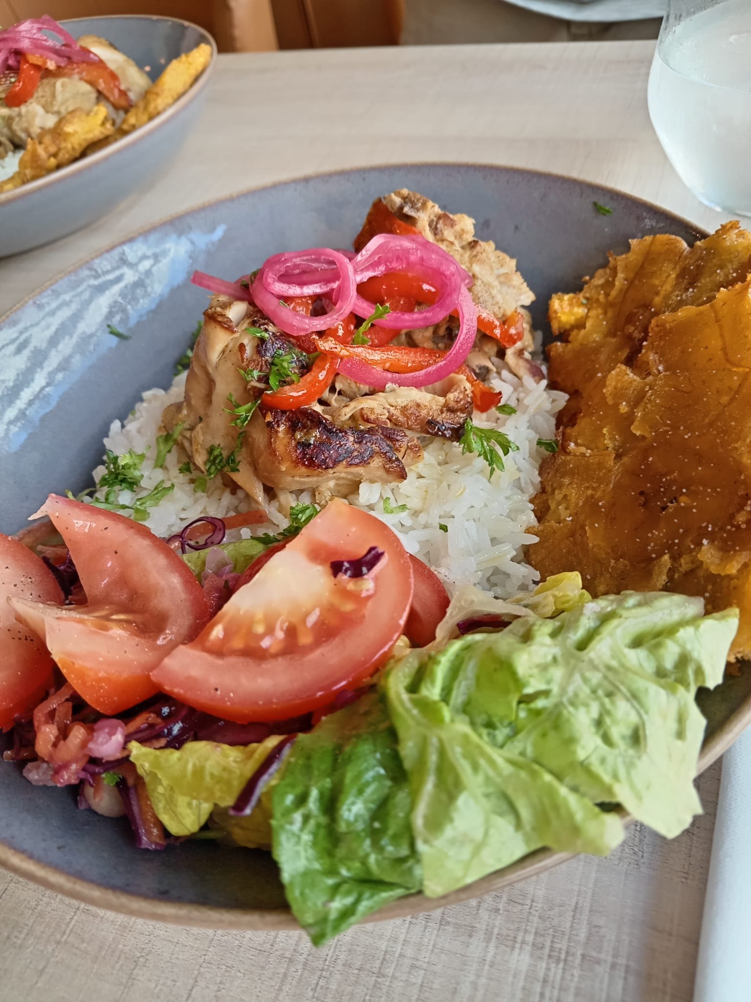 A plate of white rice topped with grilled chicken, red peppers, pickled red onions, and fresh herbs, served with a side of fried plantains, chopped tomatoes, and lettuce.